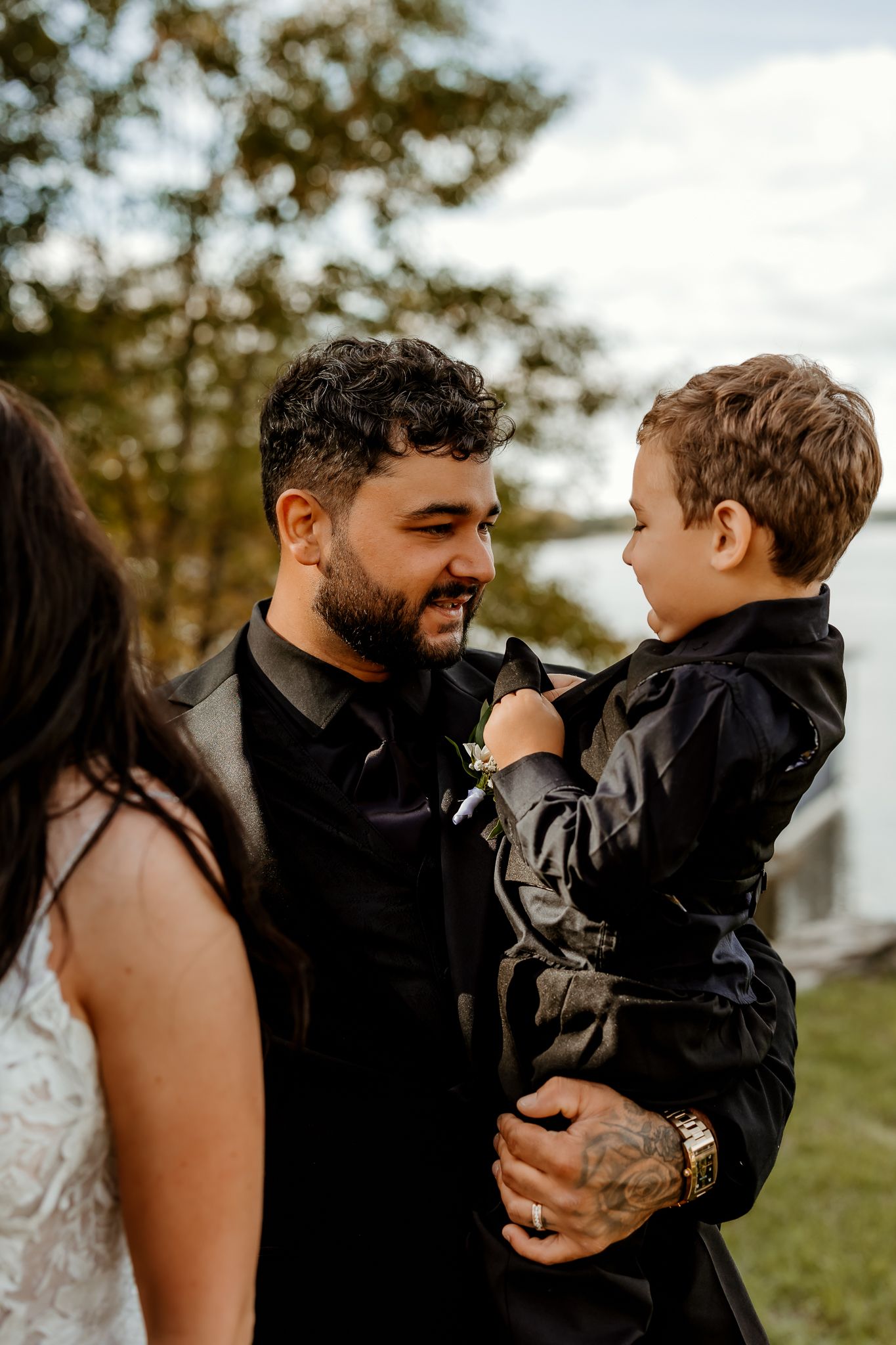 Smiling bearded man in a black shirt and jacket holds a young boy in a matching black outfit beside a woman in a lace dress outdoors.