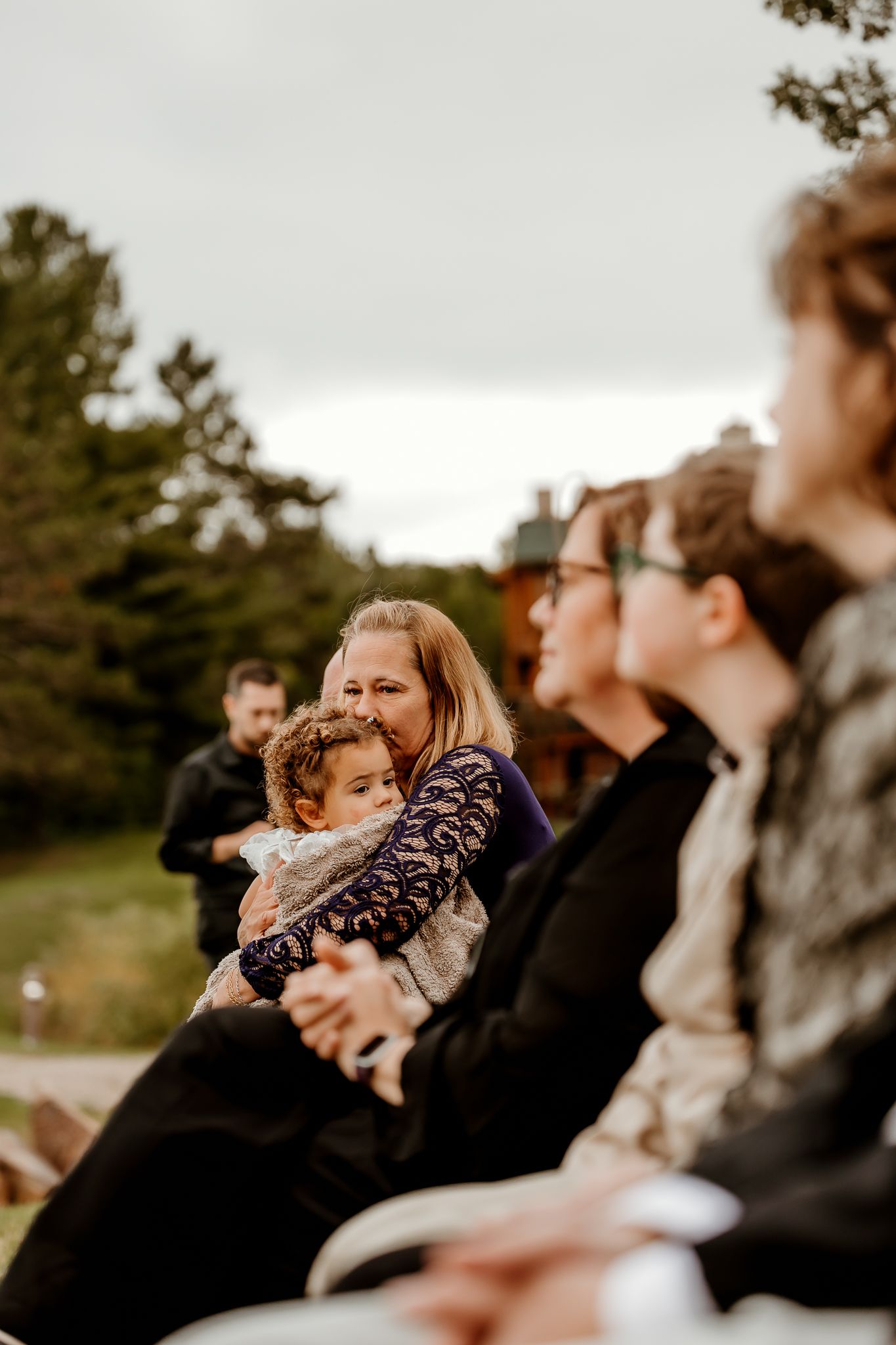 Woman in a lace-sleeved dress holds a toddler on her lap while seated among guests at an outdoor ceremony with trees in the background.