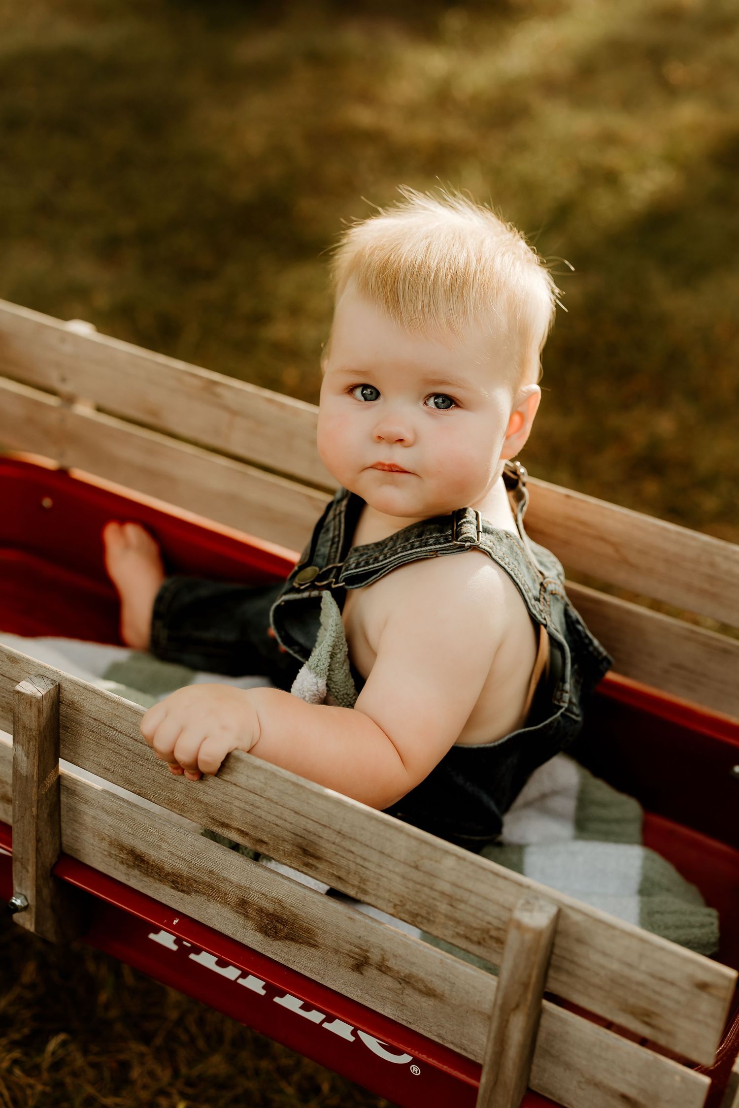 Adorable blonde toddler in denim overalls sitting in a red wooden wagon outdoors.