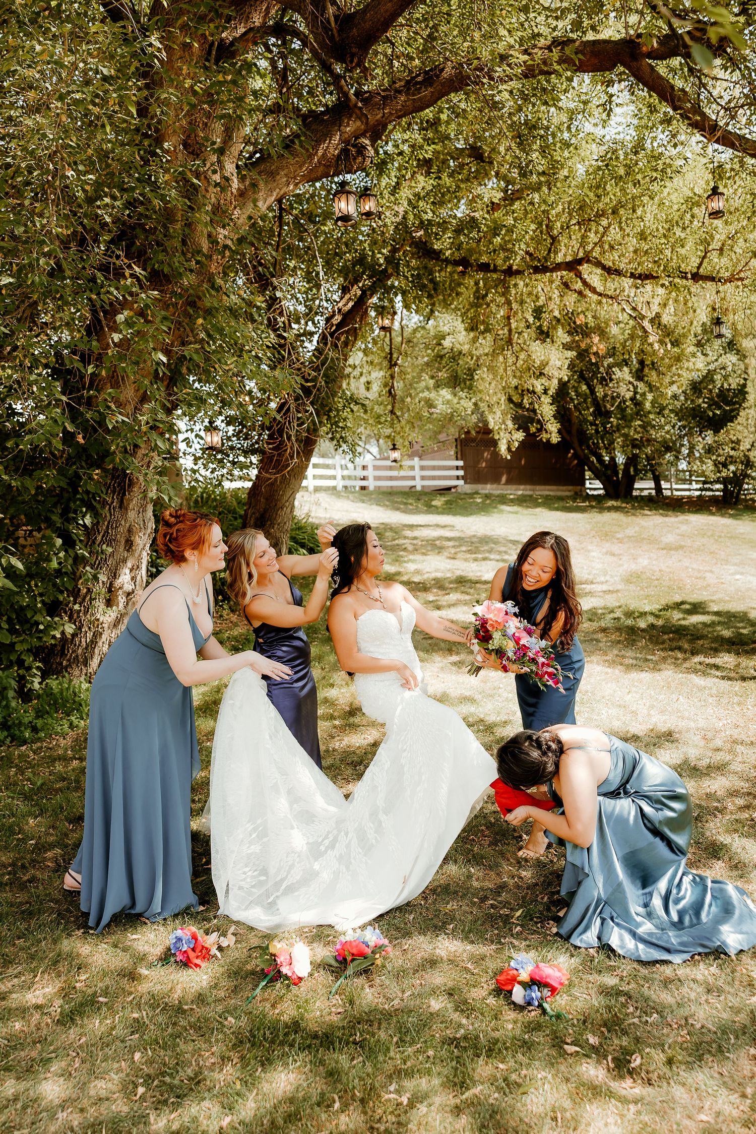 Bridesmaids in dusty blue dresses help bride with her white gown under a large tree at an outdoor wedding.