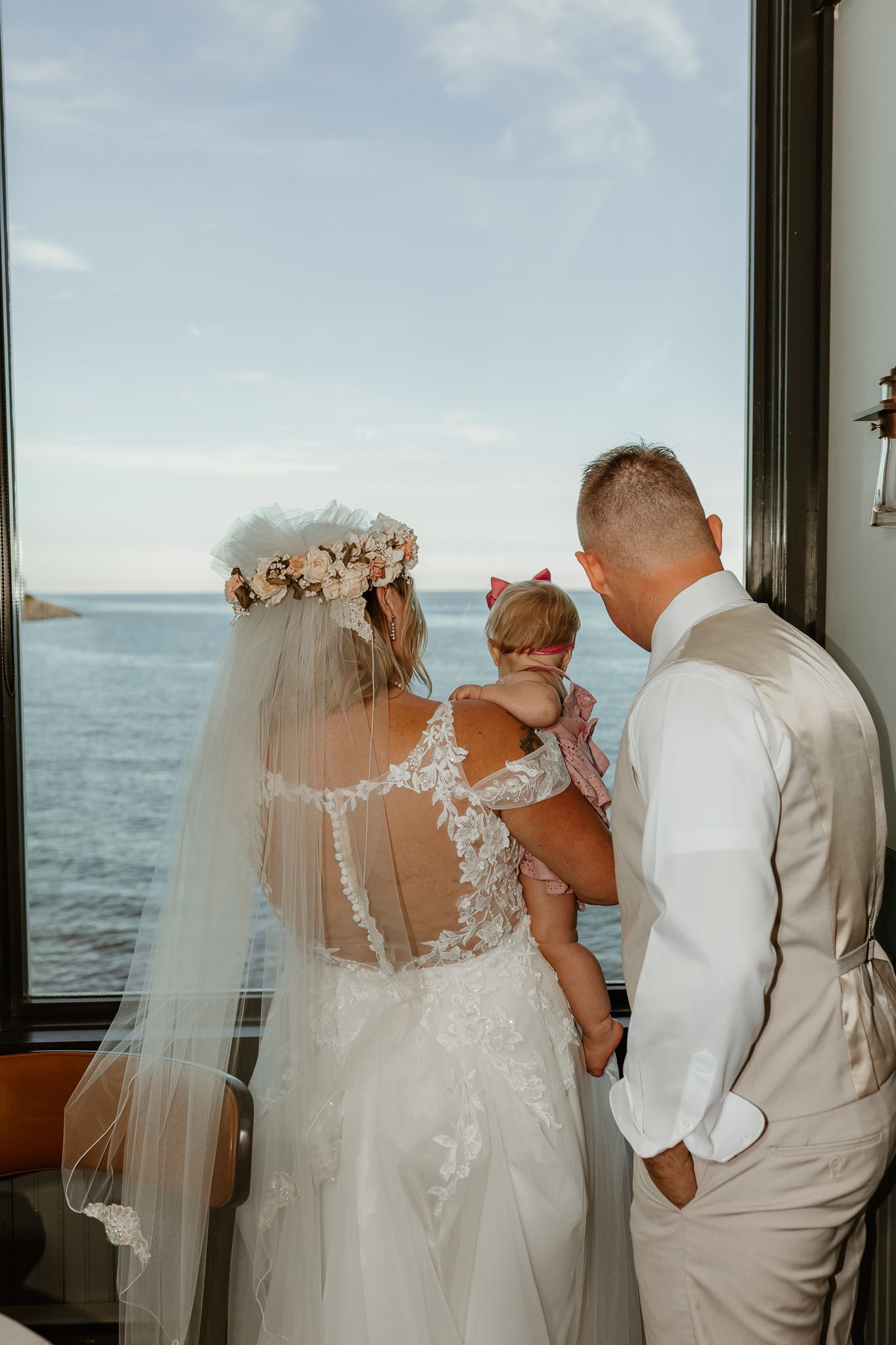 Bride in lace gown and floral crown stands with groom holding baby girl, gazing at ocean view through large window.