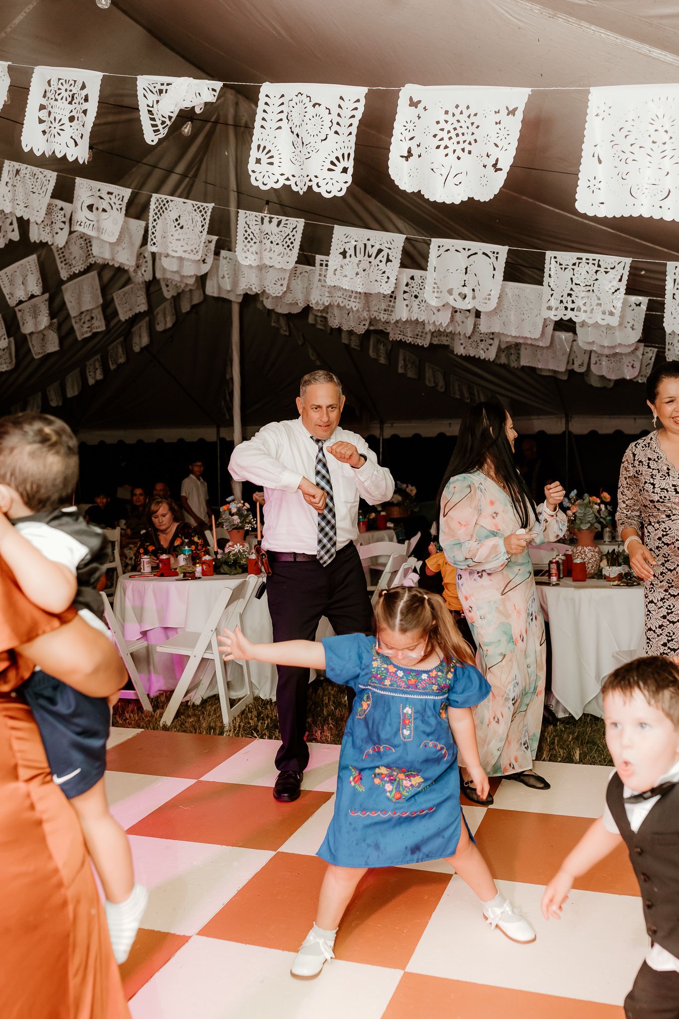 Guests dance joyfully at a festive wedding reception under a tent decorated with papel picado banners.