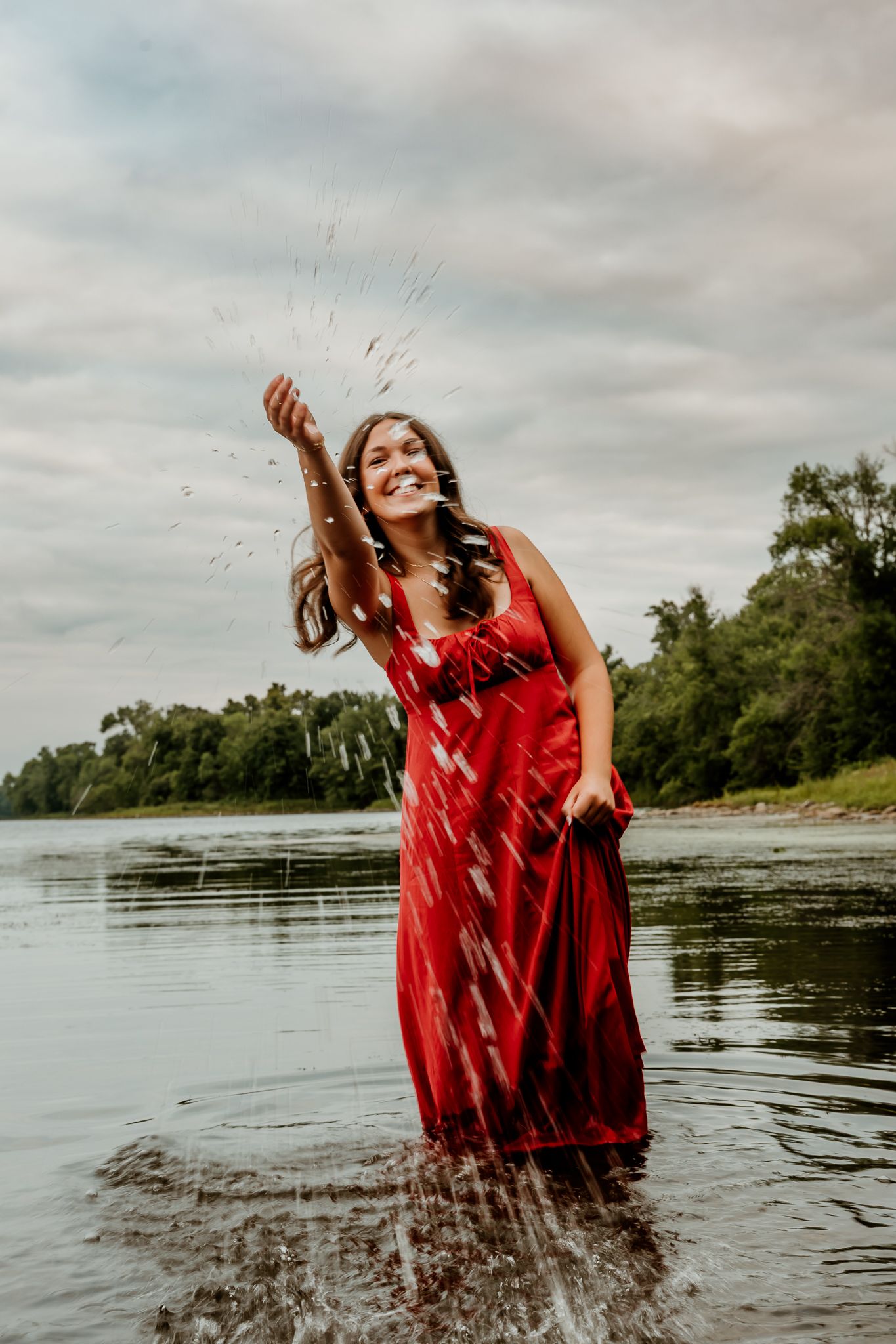 A smiling woman in a red dress splashes water while standing in a shallow river with green trees in the background.