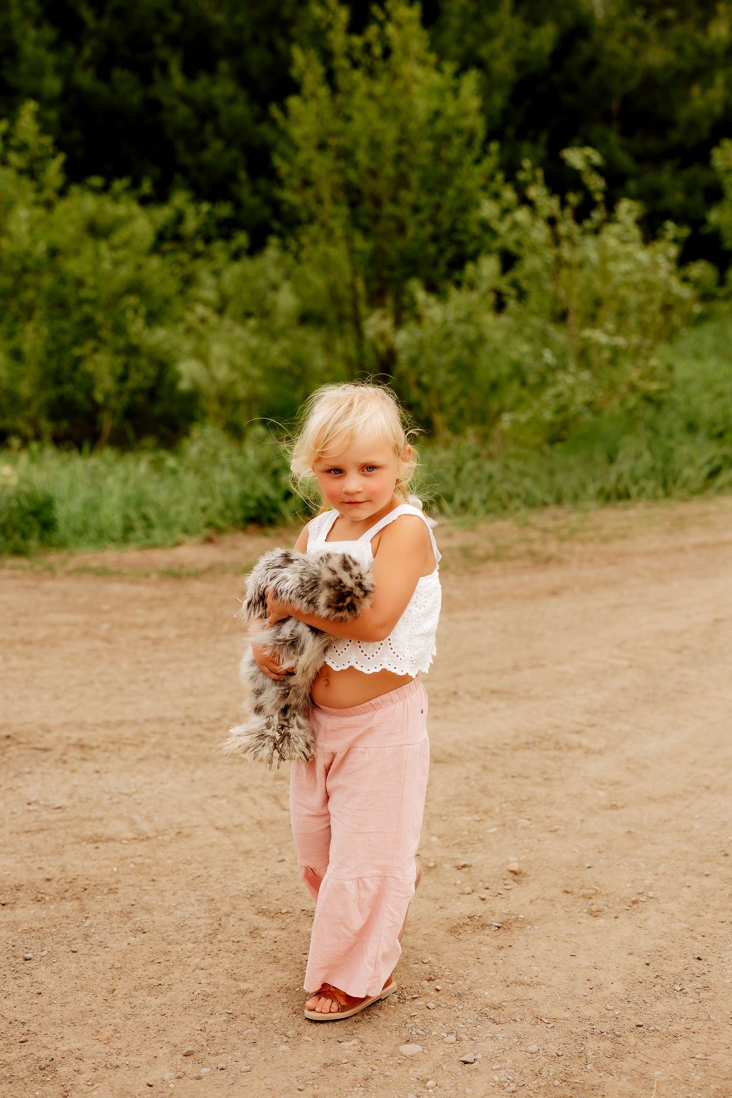 Adorable blonde toddler girl in pink pants holding a fluffy chicken on a dirt path outdoors.