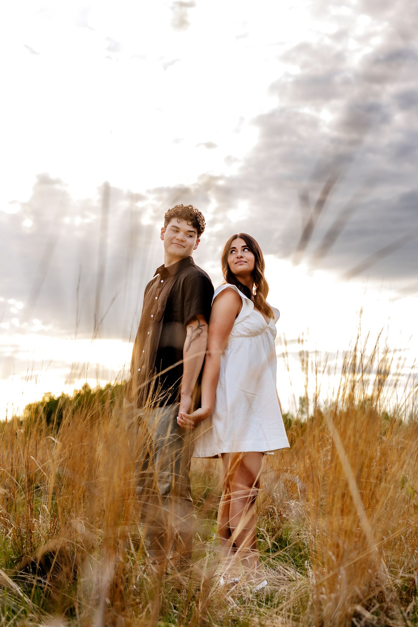 An engaged couple holds hands while standing in tall golden grass beneath a dramatic cloudy sky at sunset.