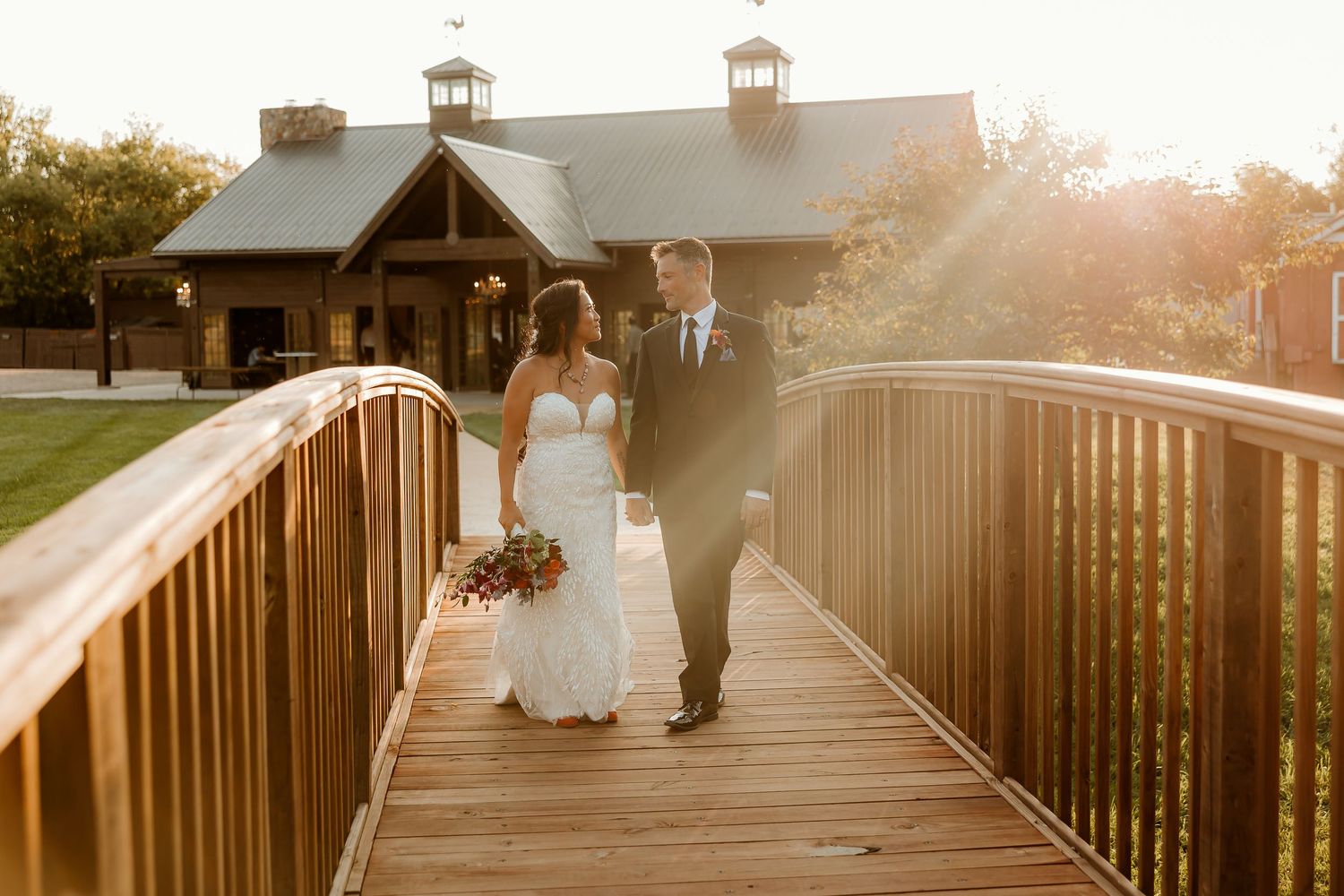 Bride and groom walk hand-in-hand on a wooden bridge at golden hour, with a rustic venue glowing in the background.