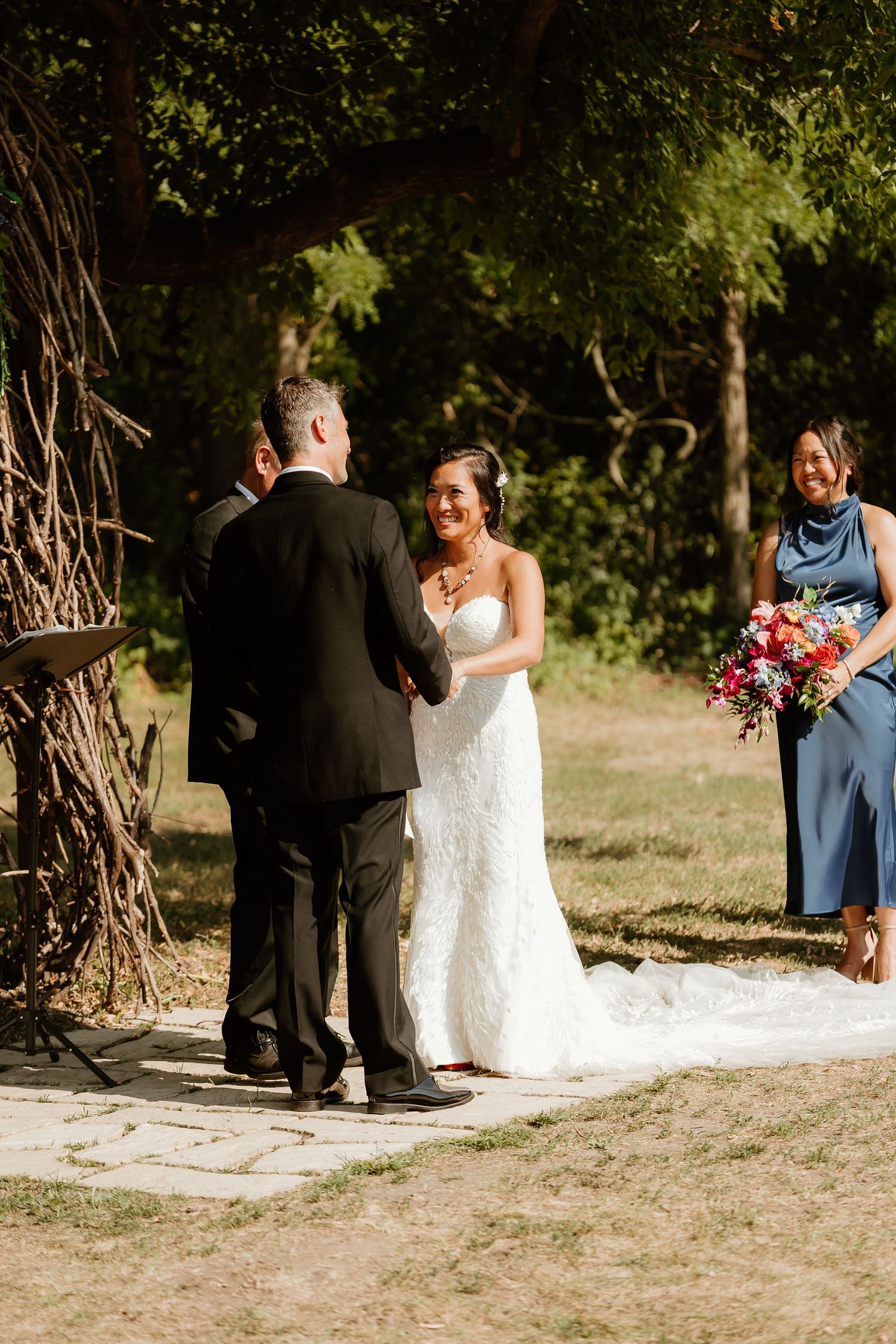 Bride in white lace gown smiles at groom during outdoor wedding ceremony with bridesmaid holding colorful bouquet.