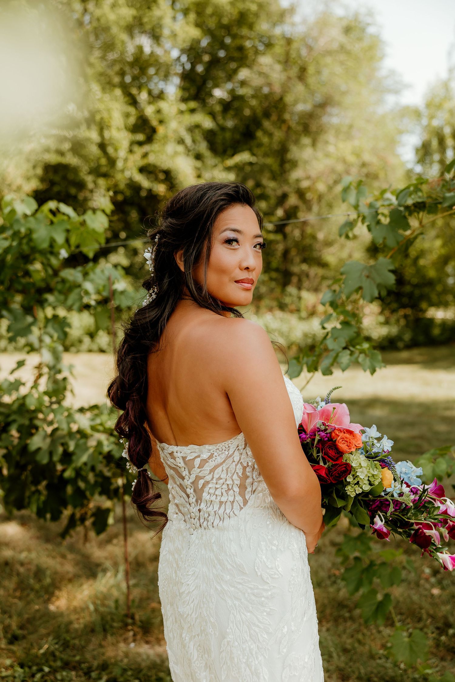 Bride in lace backless wedding gown holds colorful bouquet, posing outdoors among lush green trees.