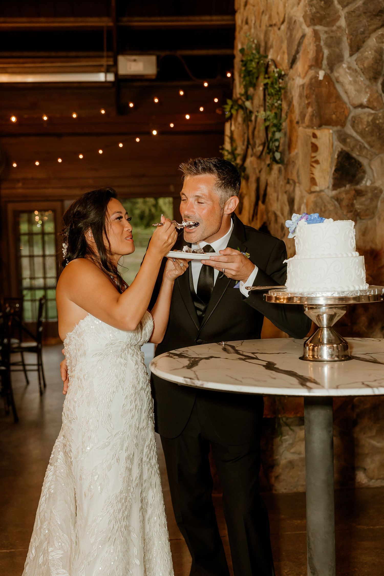Bride and groom feeding each other wedding cake at rustic stone venue with warm string lights.