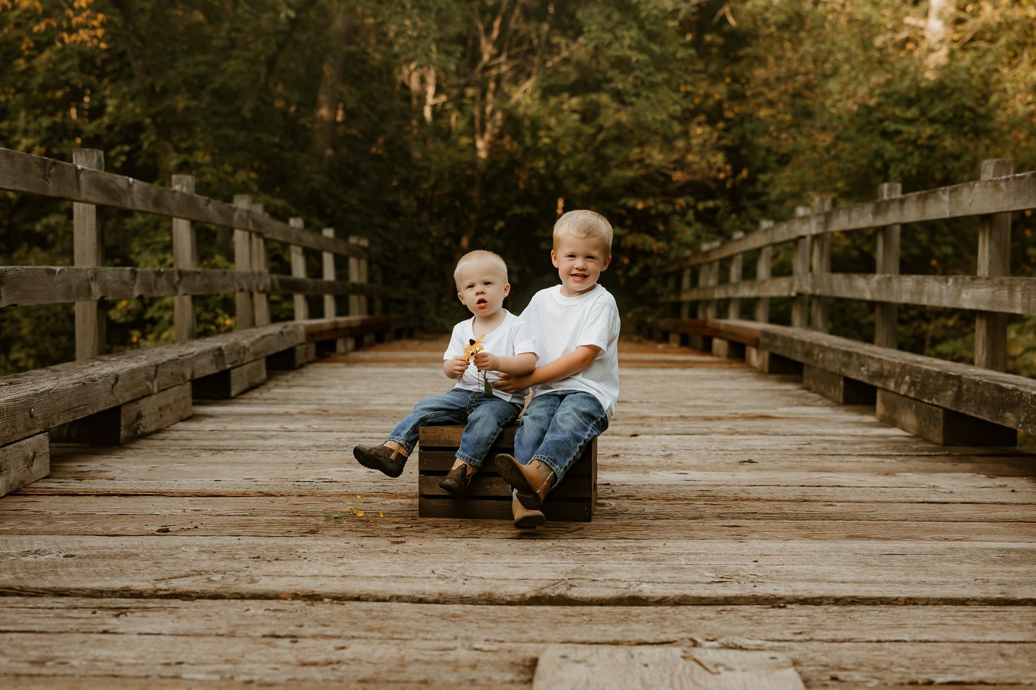Two young boys sitting together on a wooden bridge surrounded by lush green trees, smiling outdoors.