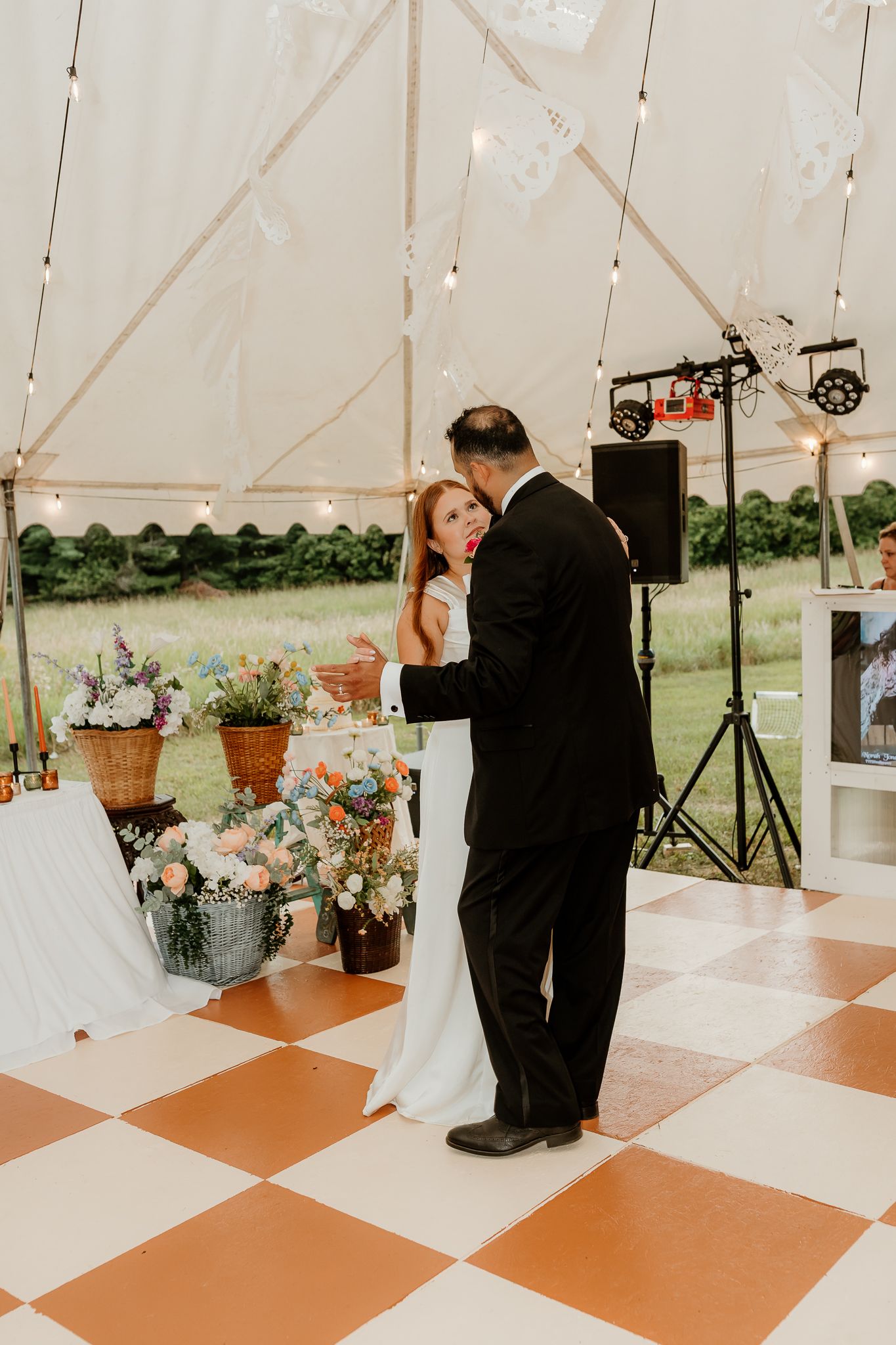 Couple sharing first dance on checkered floor under a tent with string lights and floral arrangements at an outdoor wedding.