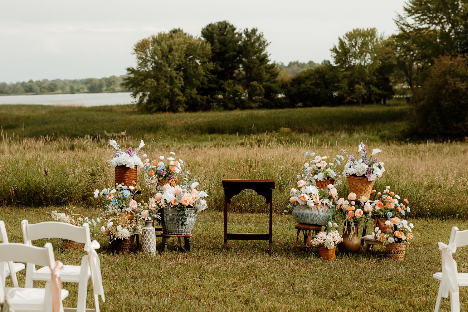 Outdoor wedding ceremony setup with floral arrangements in pastel tones, set against a scenic meadow and lake backdrop.