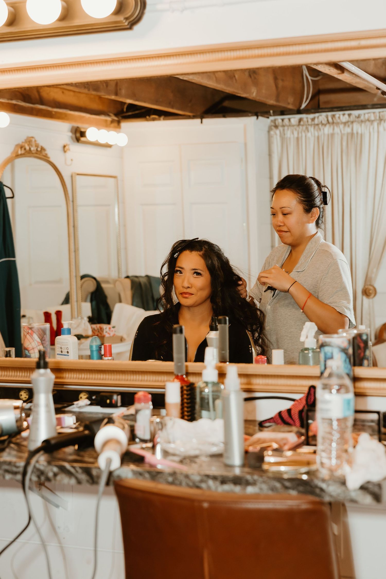 A hairstylist curls a woman's dark hair at a vanity mirror surrounded by beauty products.
