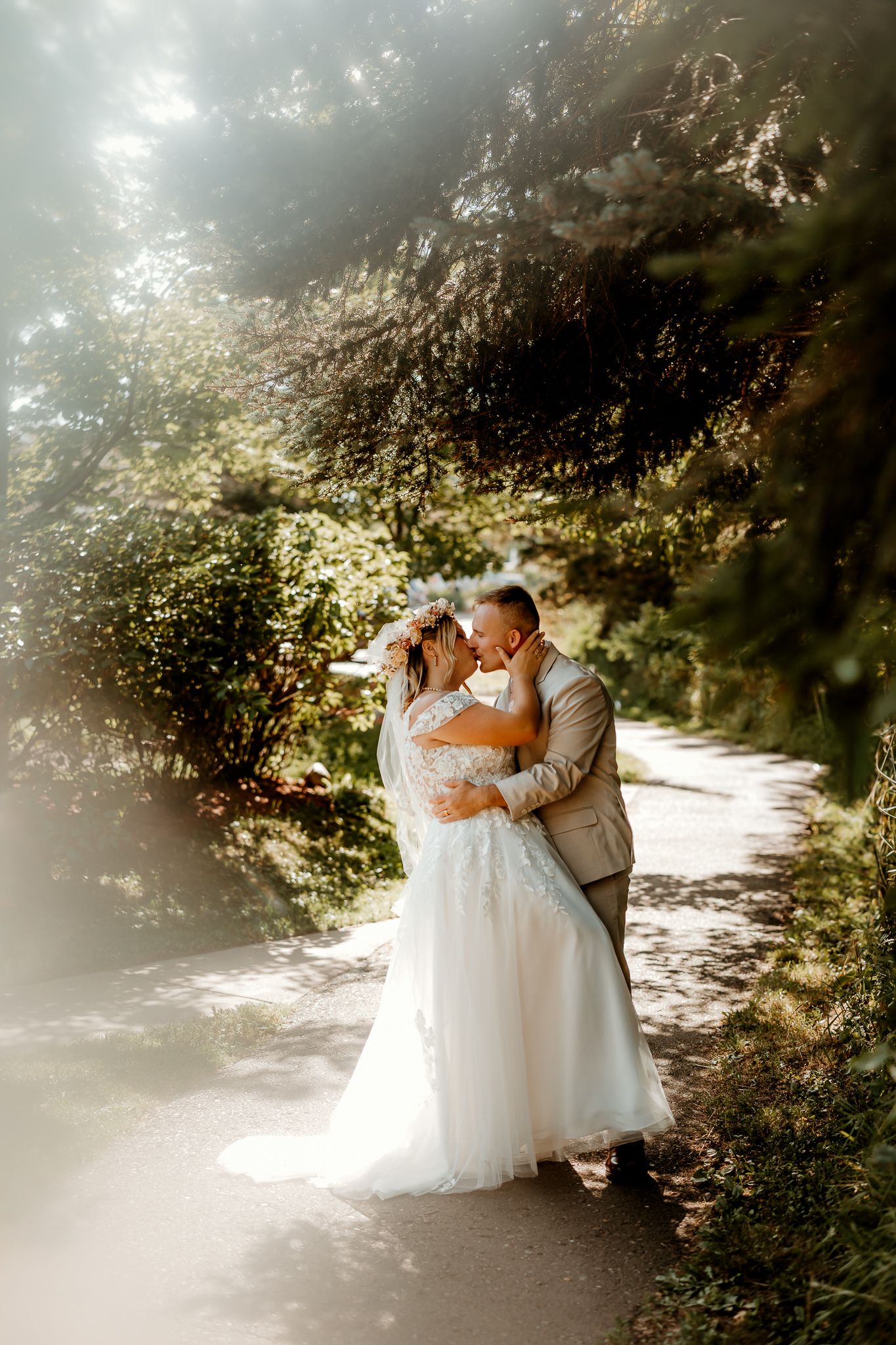 A bride and groom share a romantic embrace on a sunlit garden path surrounded by lush green trees.