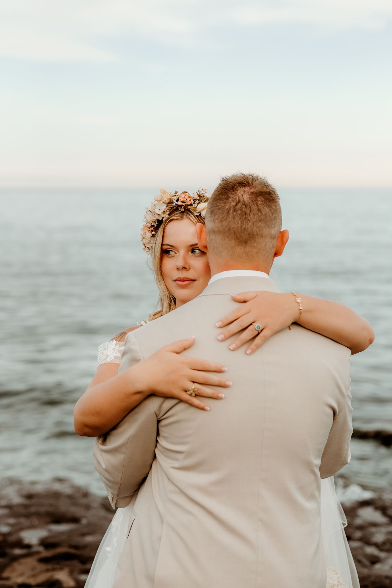 Bride in floral crown embraces groom by the ocean at sunset during a romantic beach wedding ceremony.