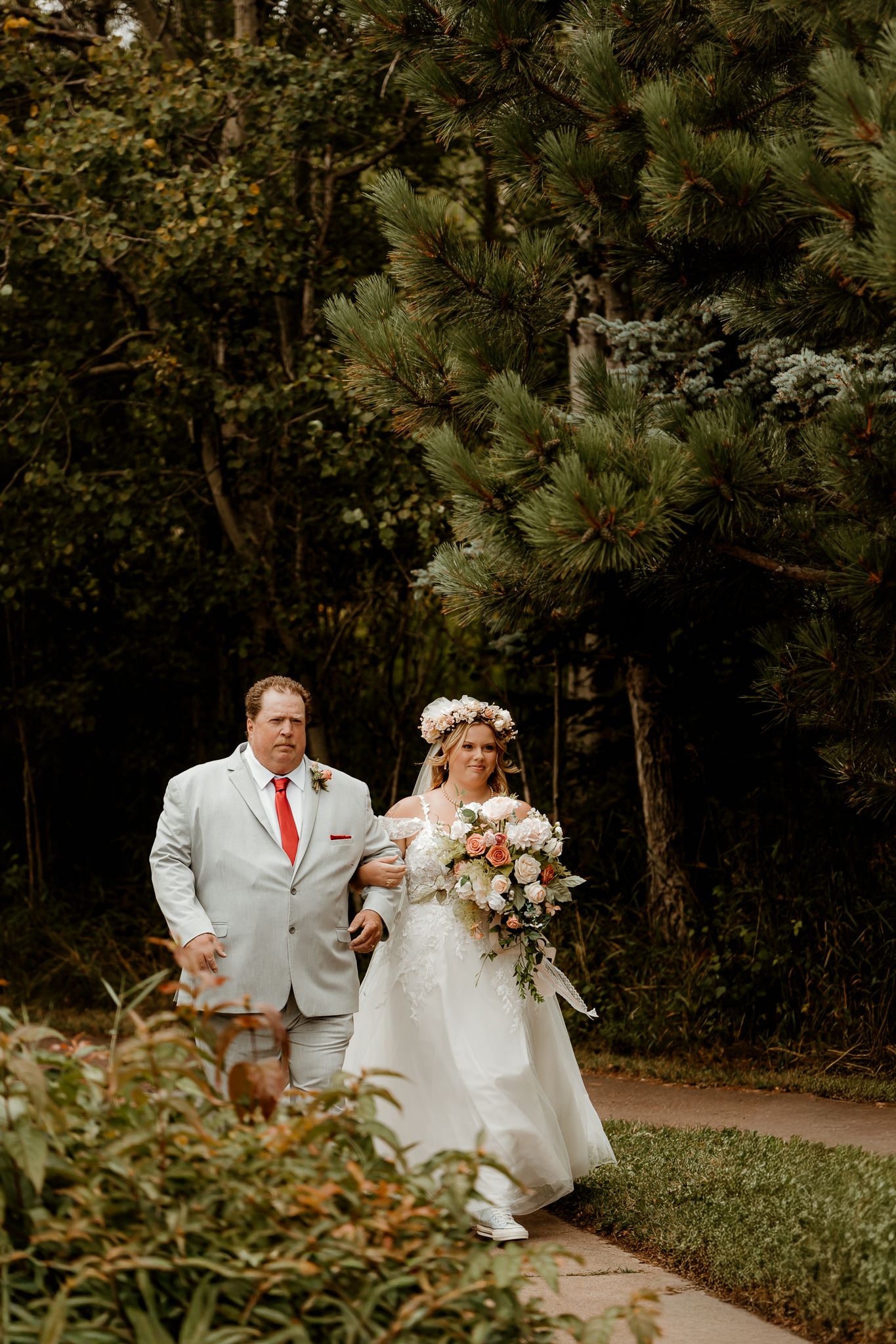 Bride in floral crown holding bouquet walks with man in gray suit on garden path surrounded by lush green trees.