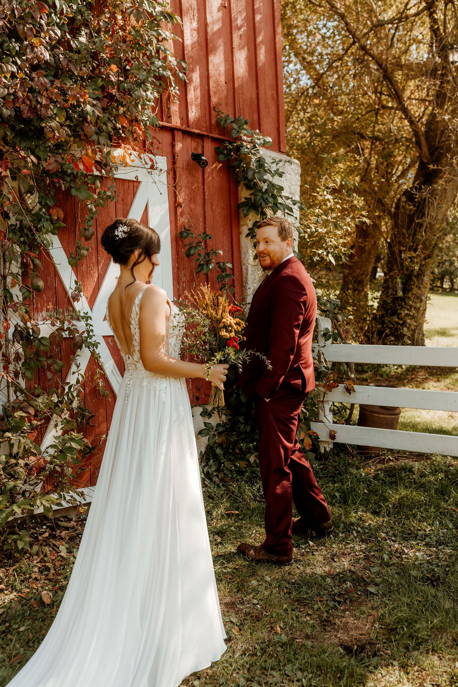 Bride in a flowing white gown holds an autumn bouquet while smiling groom in a burgundy suit looks on beside a red barn.