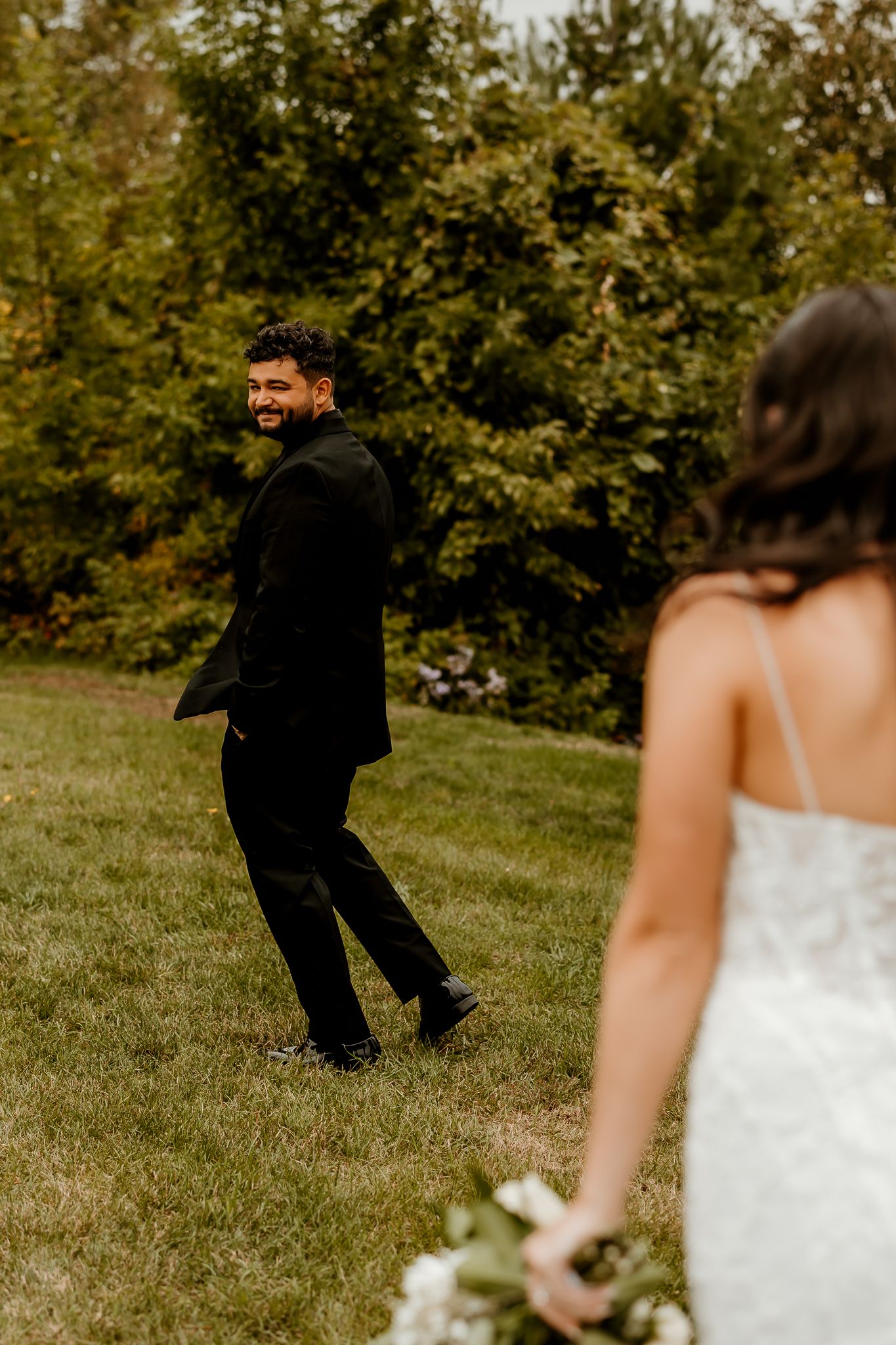 Smiling groom in a black suit walks across a sunny grassy lawn toward a bride in a lace gown holding a white bouquet.