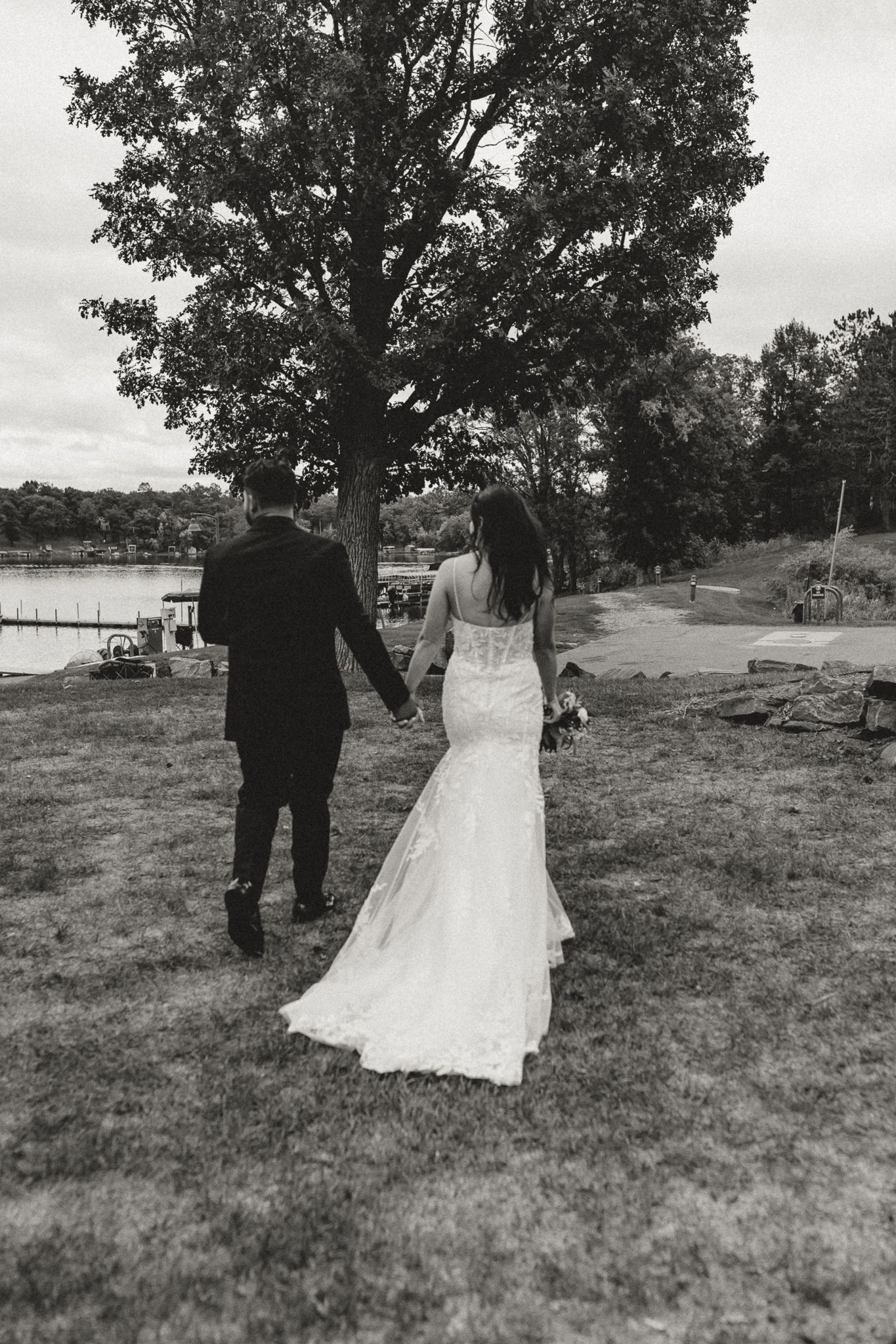 Bride in a lace wedding gown and groom in a dark suit hold hands as they walk across a grassy lawn toward a lake in black and white.