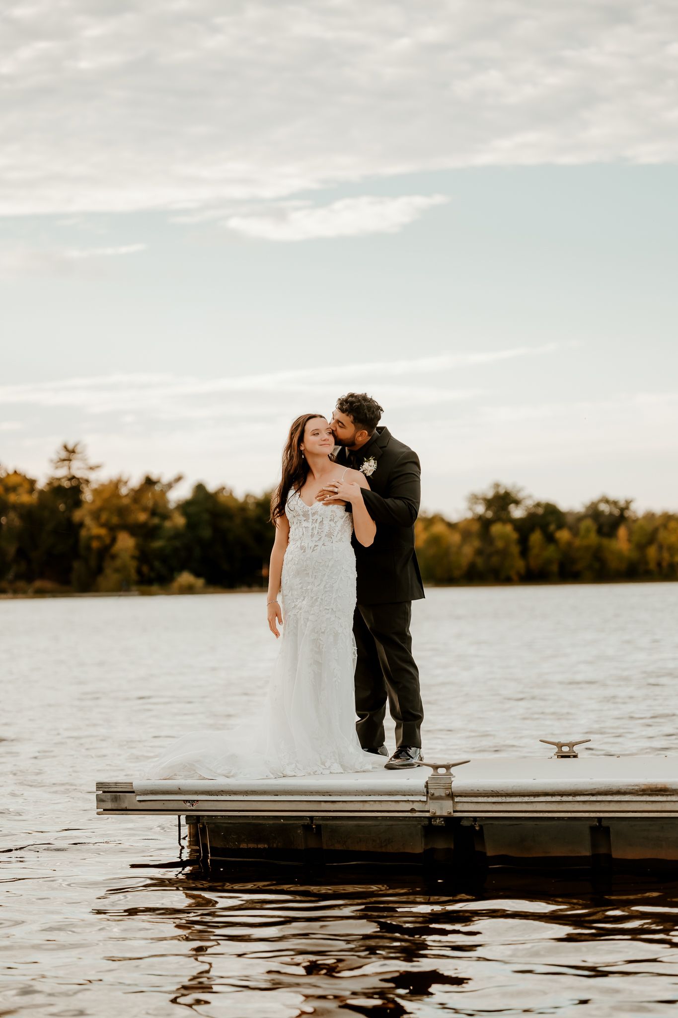 Bride in a lace wedding gown and groom in a dark suit embrace on a lakeside dock at sunset, with autumn trees in the background.