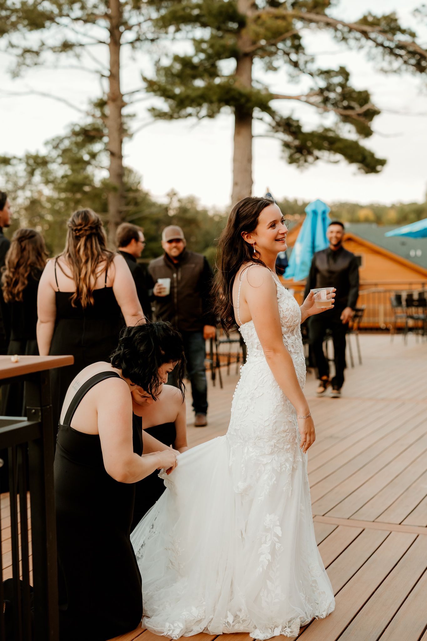 Smiling bride in a lace wedding gown holds a drink on a wooden deck while a bridesmaid in black adjusts her train and guests mingle nearby.