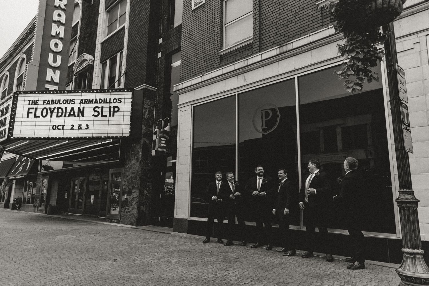Groomsmen in dark suits stand in a row on a city sidewalk beside a brick building and a theater marquee reading The Fabulous Armadillos.