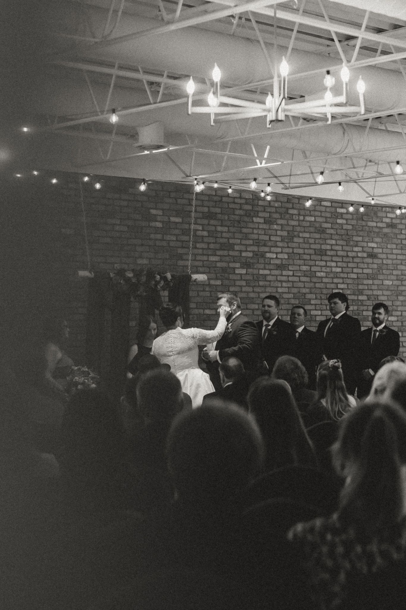 Black and white photo of a bride and groom at the altar during a wedding ceremony in a brick venue with chandeliers and string lights.