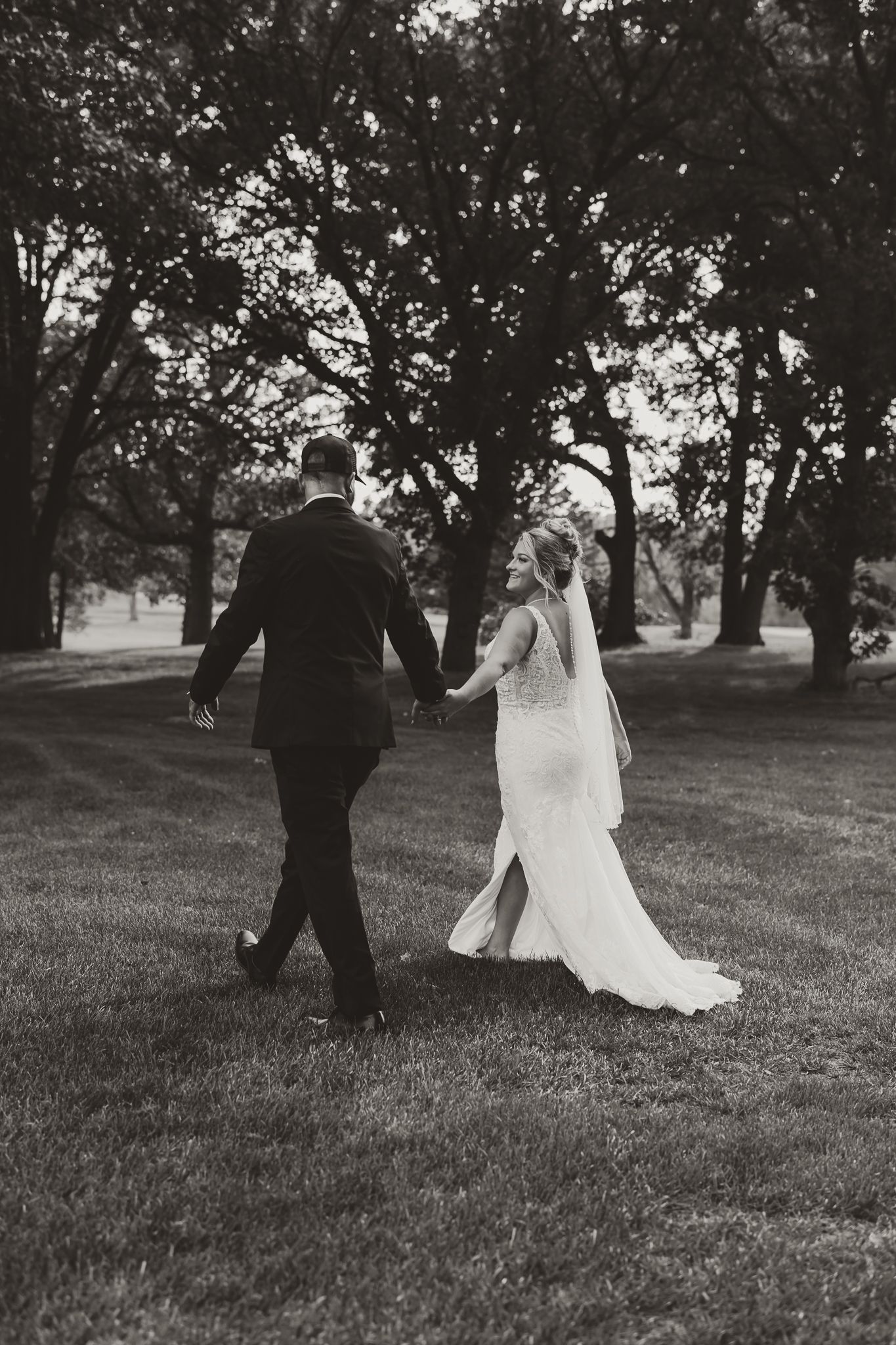 Black and white photo of a bride and groom holding hands walking through a tree-lined lawn on their wedding day.