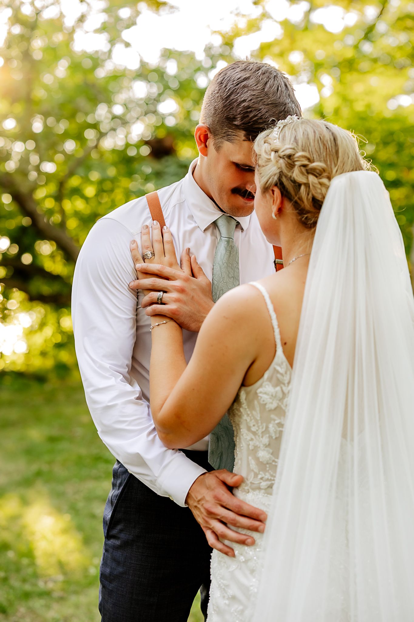 Bride and groom share an intimate moment outdoors, with bride in lace dress and veil, groom in white shirt and tie.