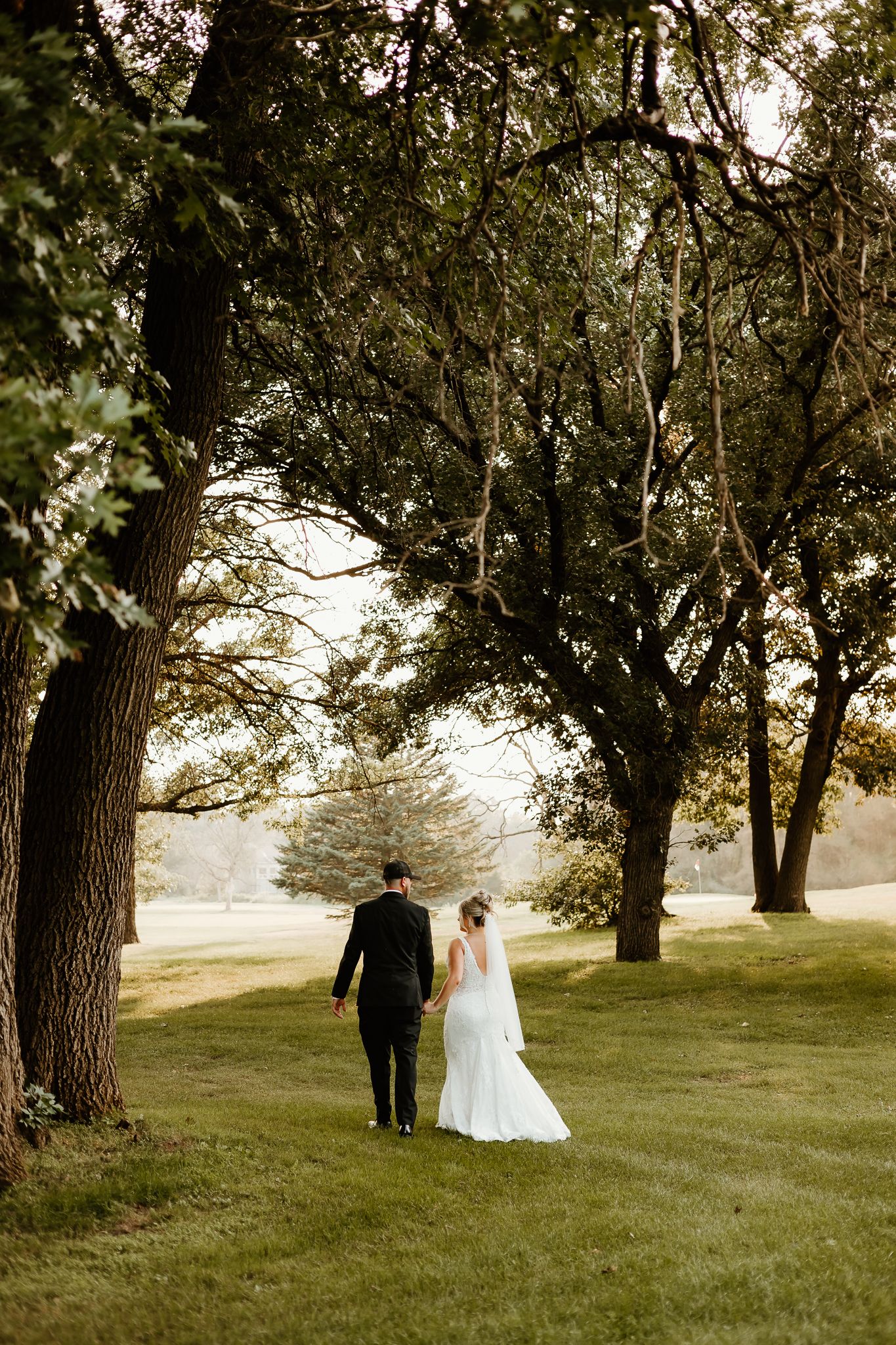 A bride and groom walk hand in hand through a lush green park surrounded by tall trees on their wedding day.