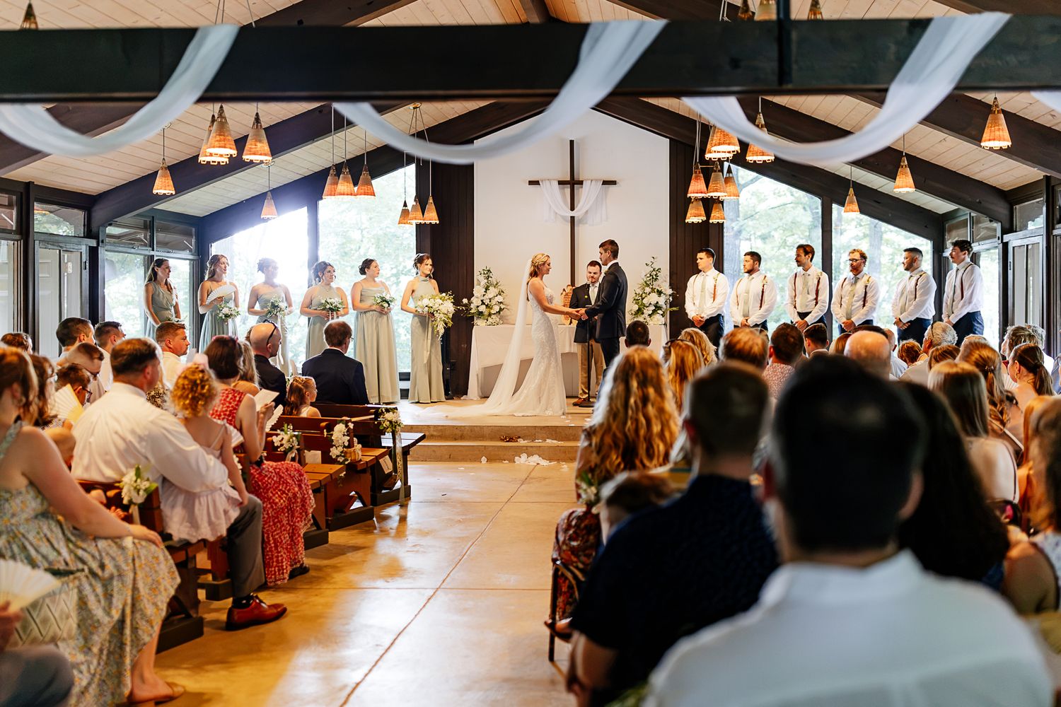 Elegant indoor wedding ceremony with bride and groom at altar, bridal party, cross, draped fabric ceiling, and seated guests.