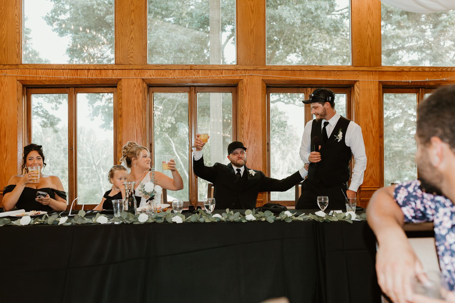 Wedding reception toast with guests raising glasses at a decorated head table in a rustic venue with large windows.