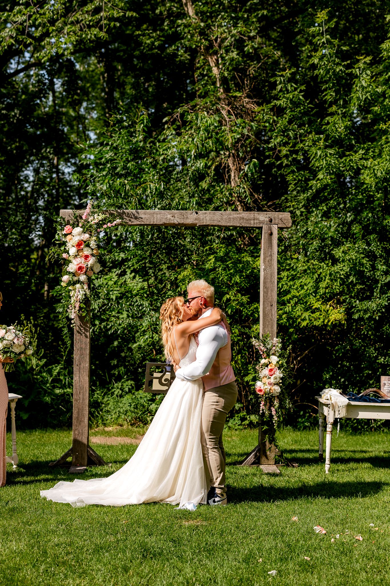 Bride and groom share first kiss under rustic wooden arch adorned with pink flowers at outdoor garden wedding ceremony.
