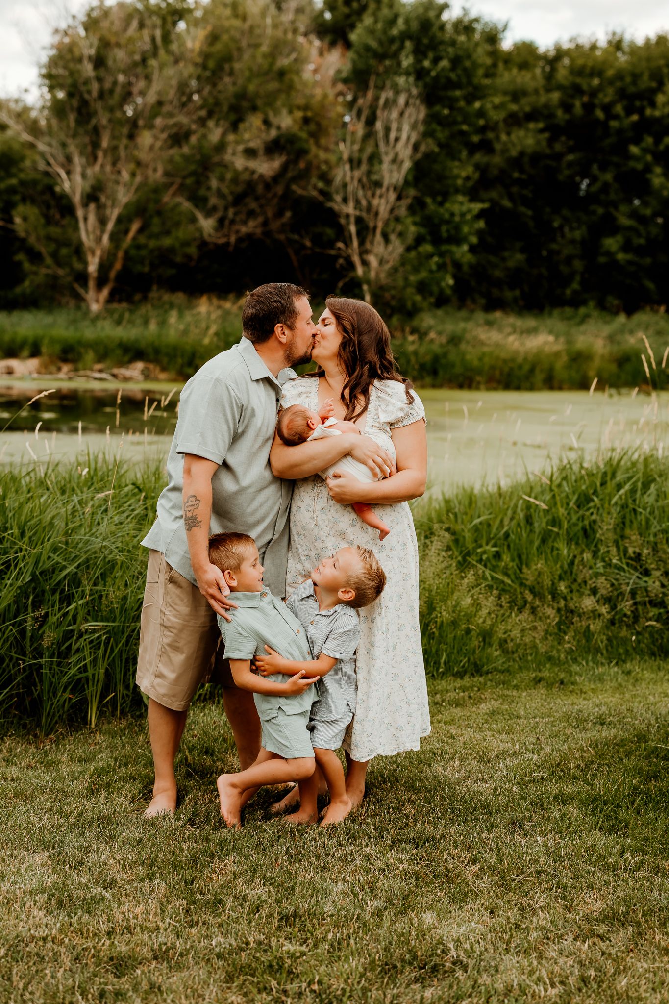 A family of five poses outdoors near a pond, parents kissing while holding a newborn, two young boys standing in front.