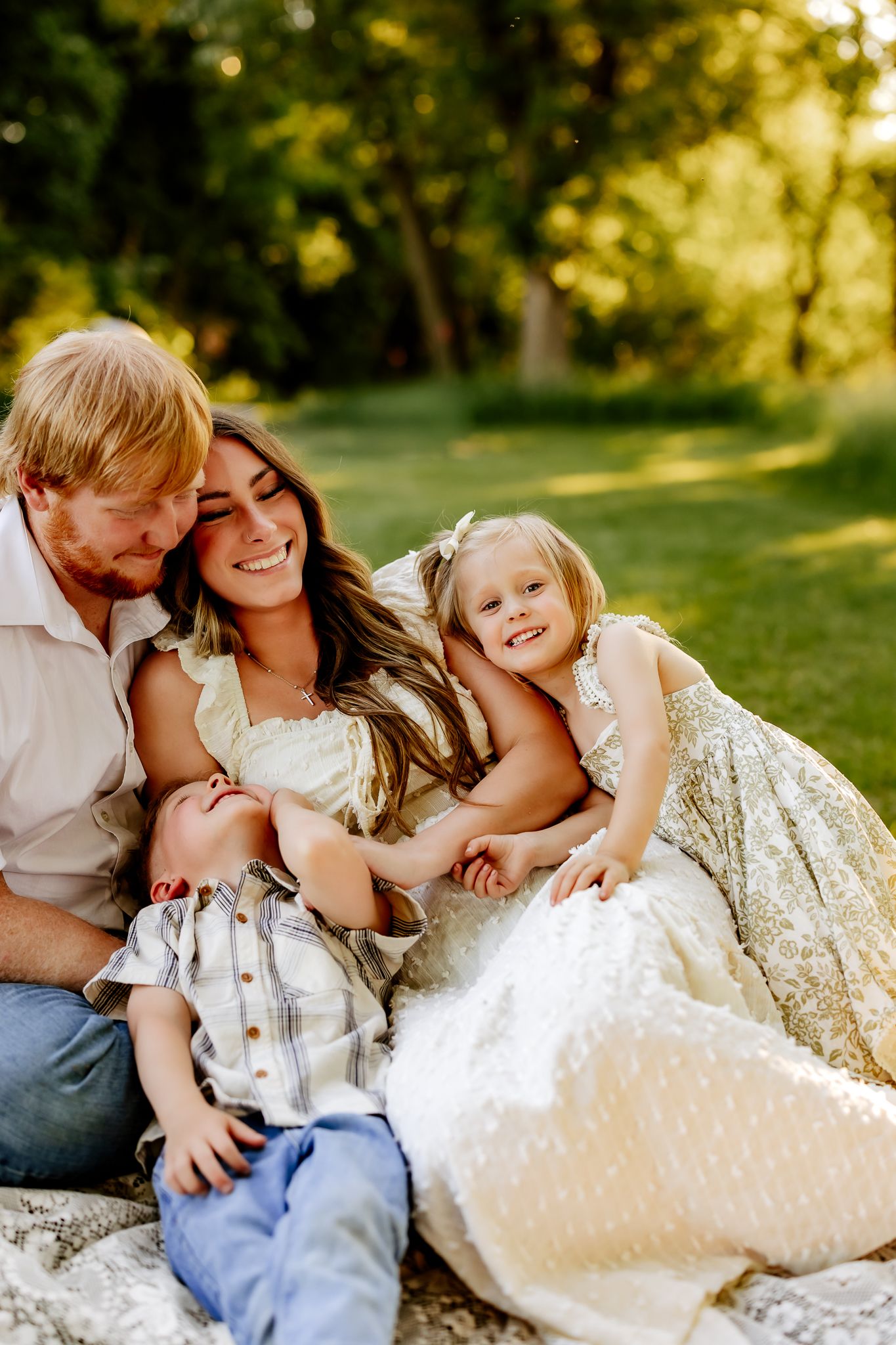 A cheerful family of four laughs and cuddles on a white blanket in a beautiful sunlit outdoor setting.