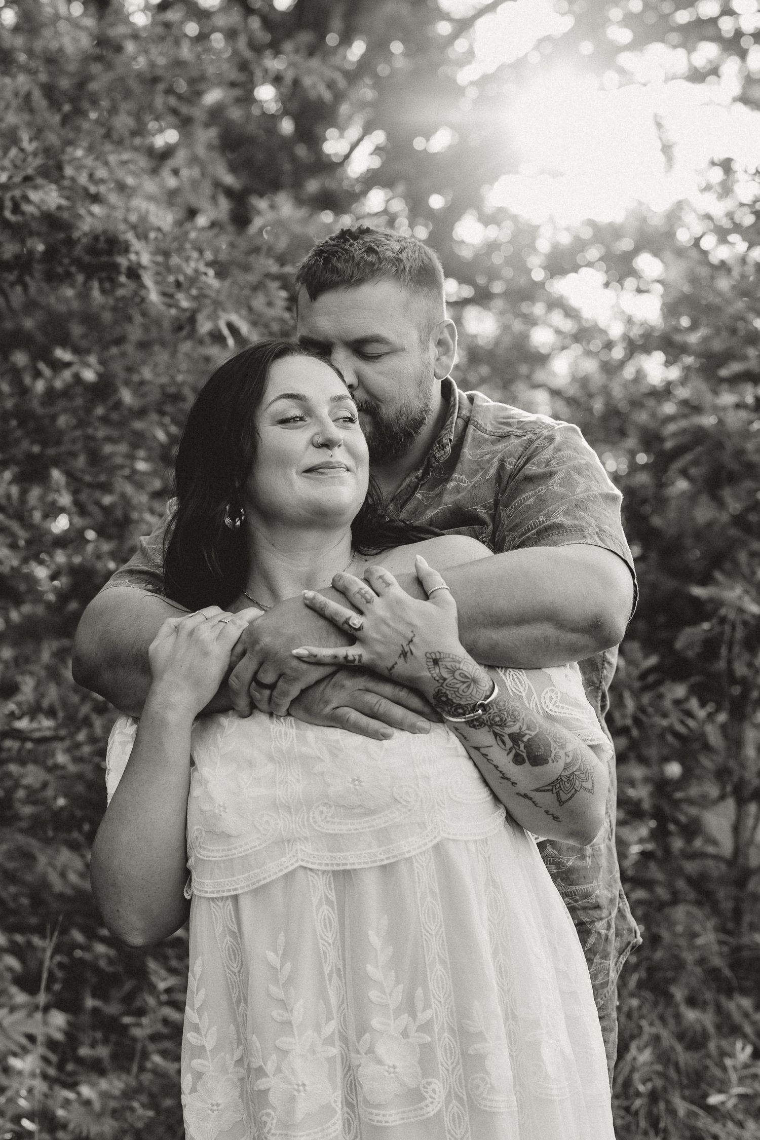 Romantic black and white maternity photo of couple embracing tenderly in sunlit garden setting.