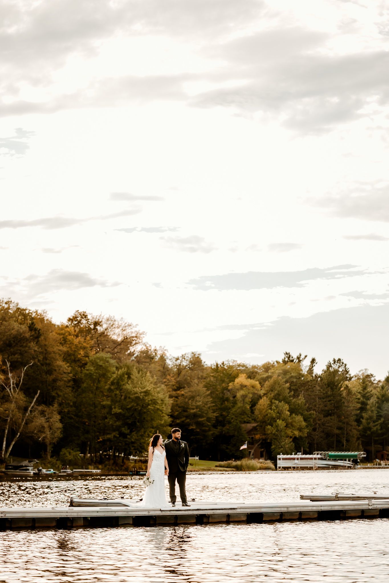 Bride and groom stand together on a lakeside wooden dock at sunset, with autumn trees and a boat across the calm water.