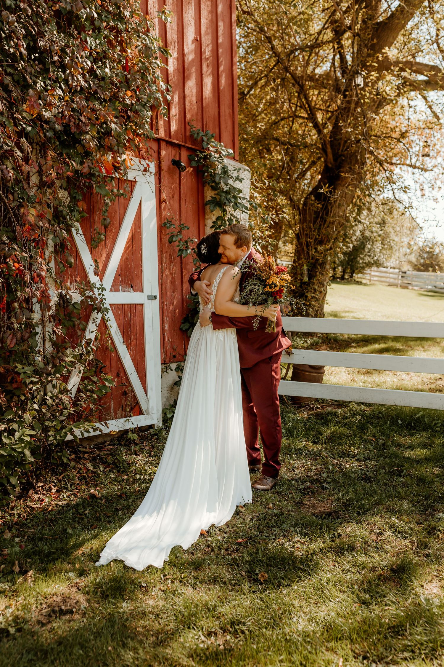 Bride in a flowing white gown and groom in a burgundy suit embrace beside a red barn with white doors and an autumn tree.