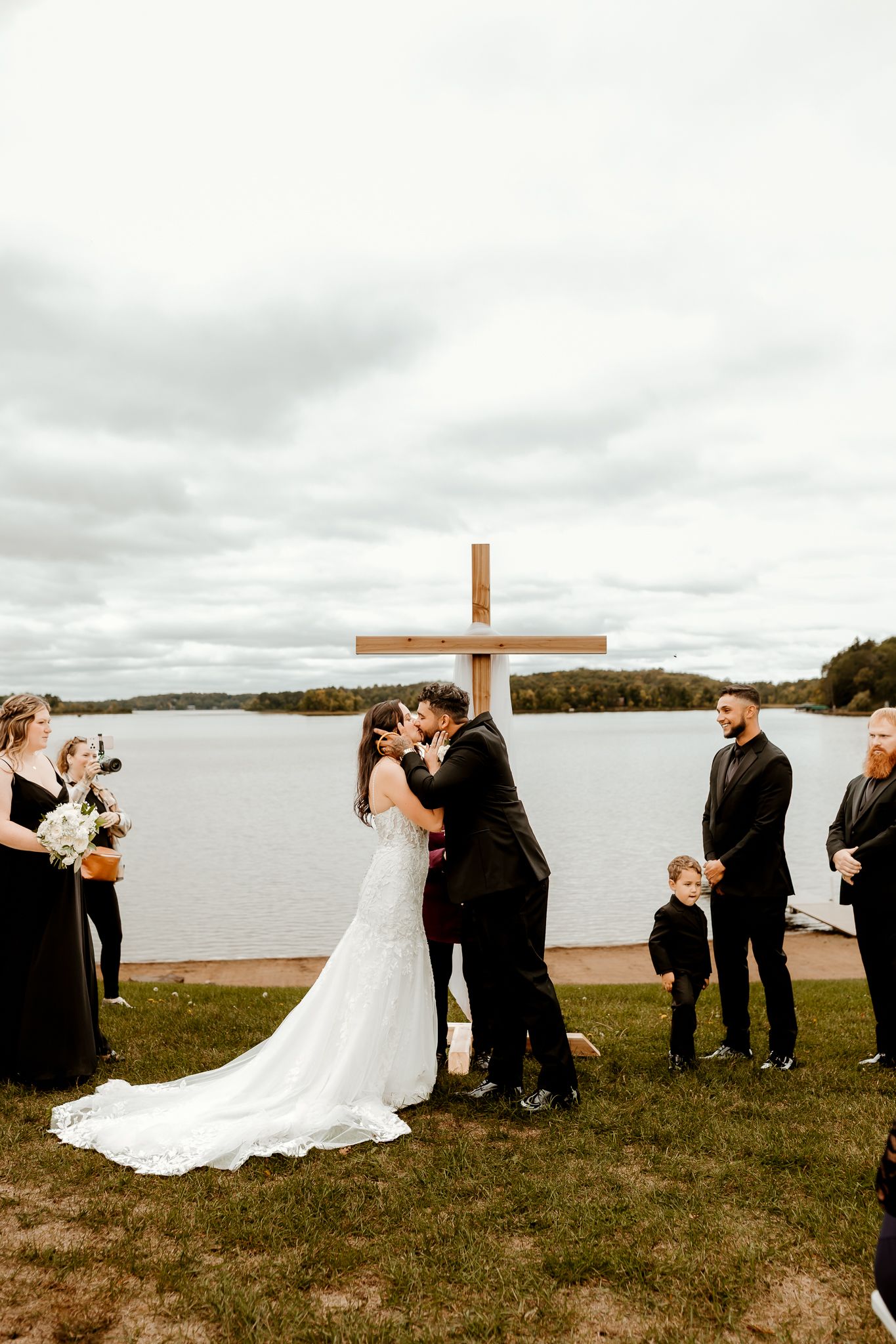 Bride in a lace gown and groom in black share a kiss at a lakeside wedding ceremony before a wooden cross, with guests looking on.