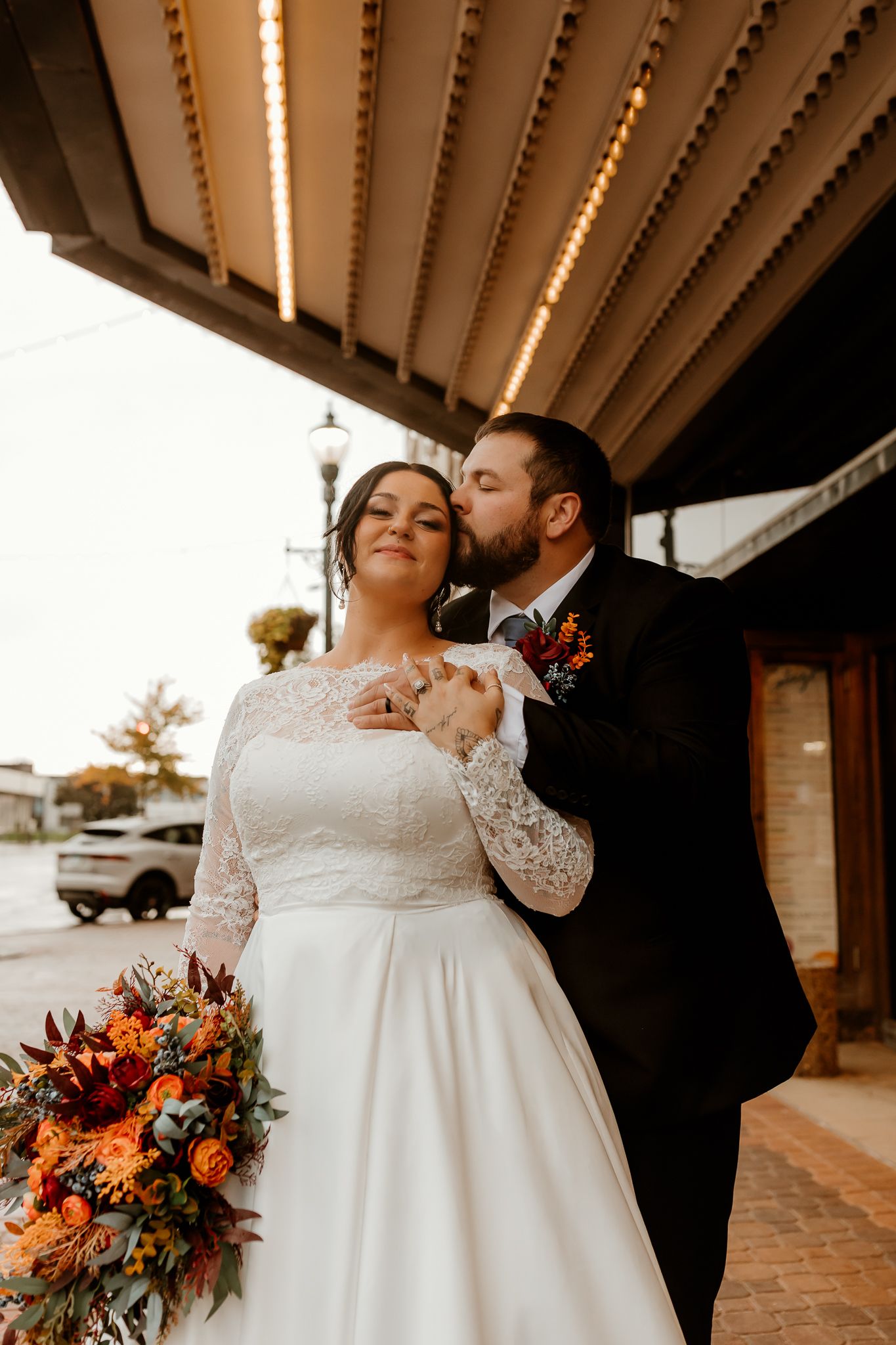 Groom in a black suit kisses his bride's forehead as she holds an orange autumn bouquet under a vintage theater marquee.