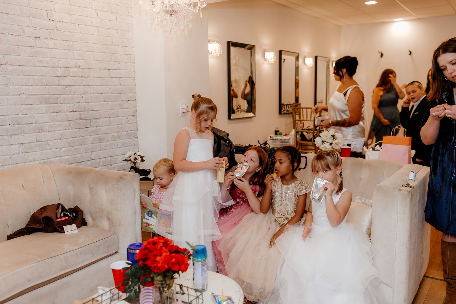 Young flower girls in white and pink dresses sit on cream sofas in a bright bridal suite while adults stand and chat nearby.