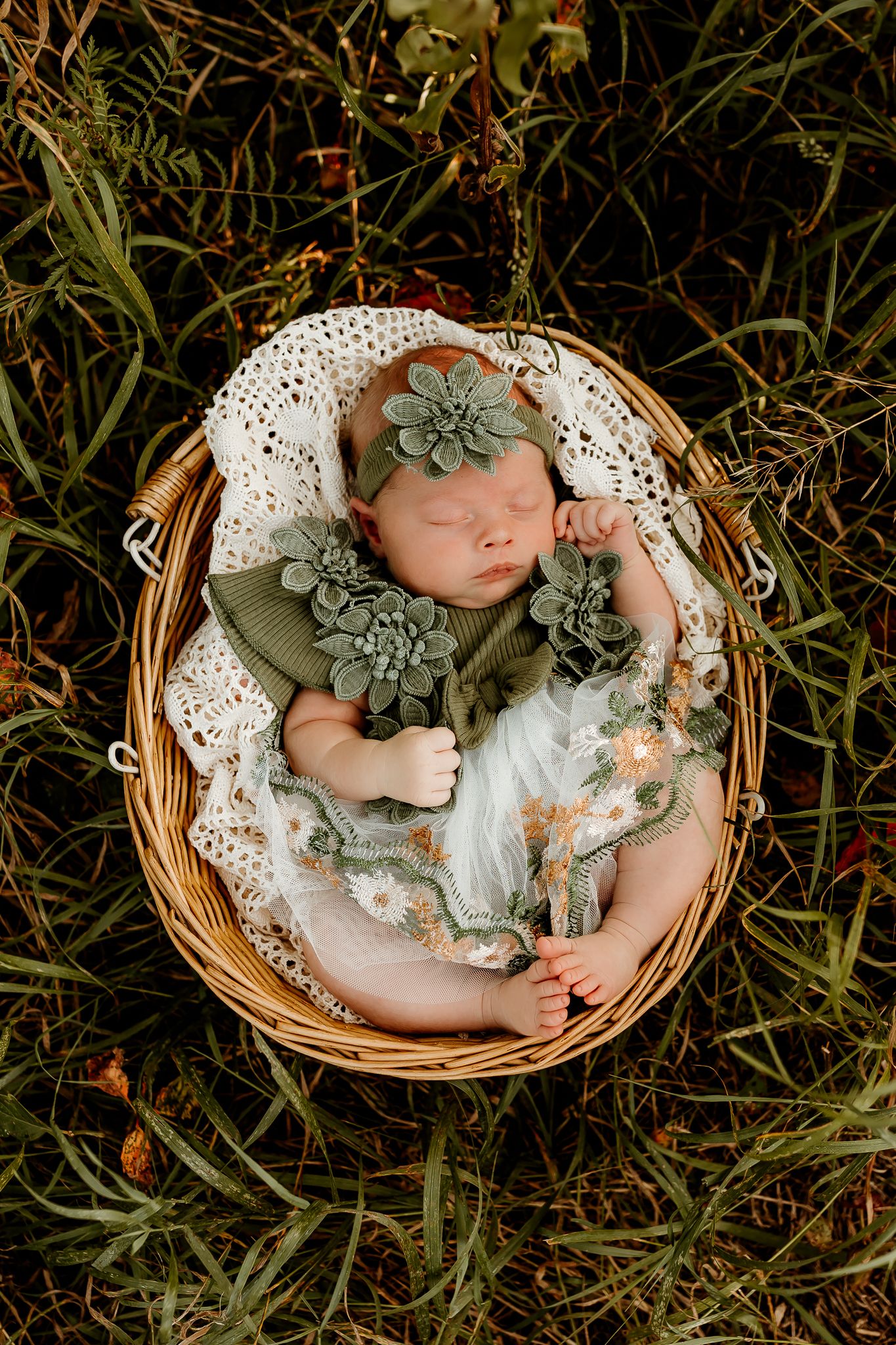 Sleeping newborn baby girl in a wicker basket wearing a green floral outfit and headband on a grass background.