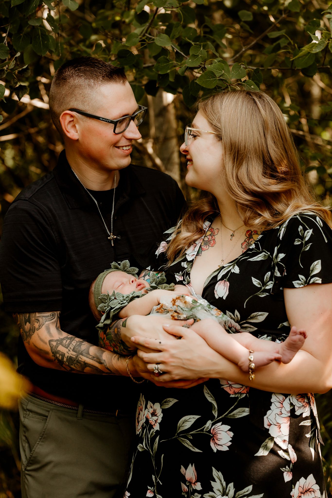 A couple smiles at each other while holding their newborn baby outdoors, man with tattoos wearing black shirt, woman in floral dress.