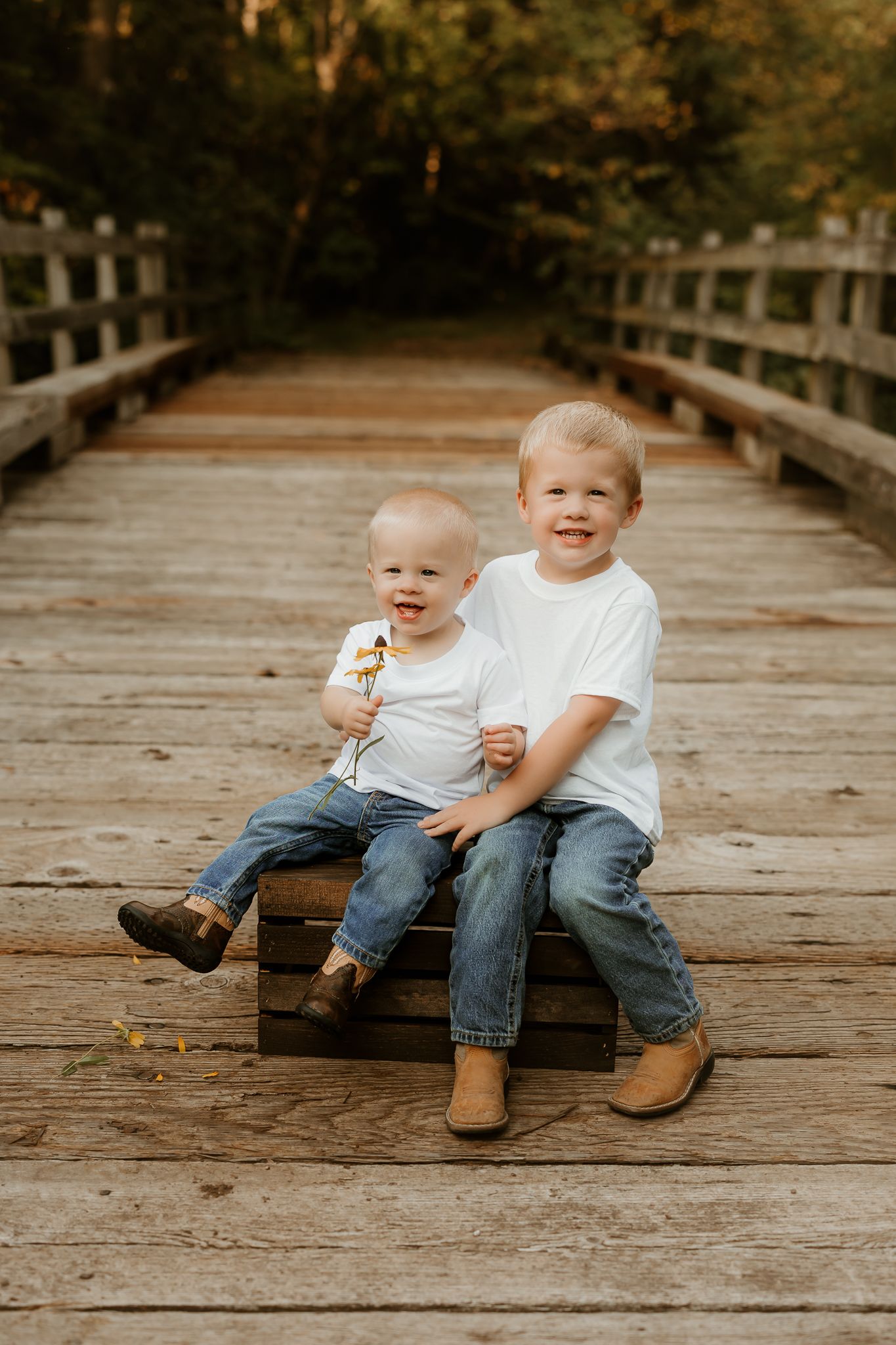 Two young blonde brothers in white shirts and jeans sit smiling on a wooden bridge surrounded by lush greenery.