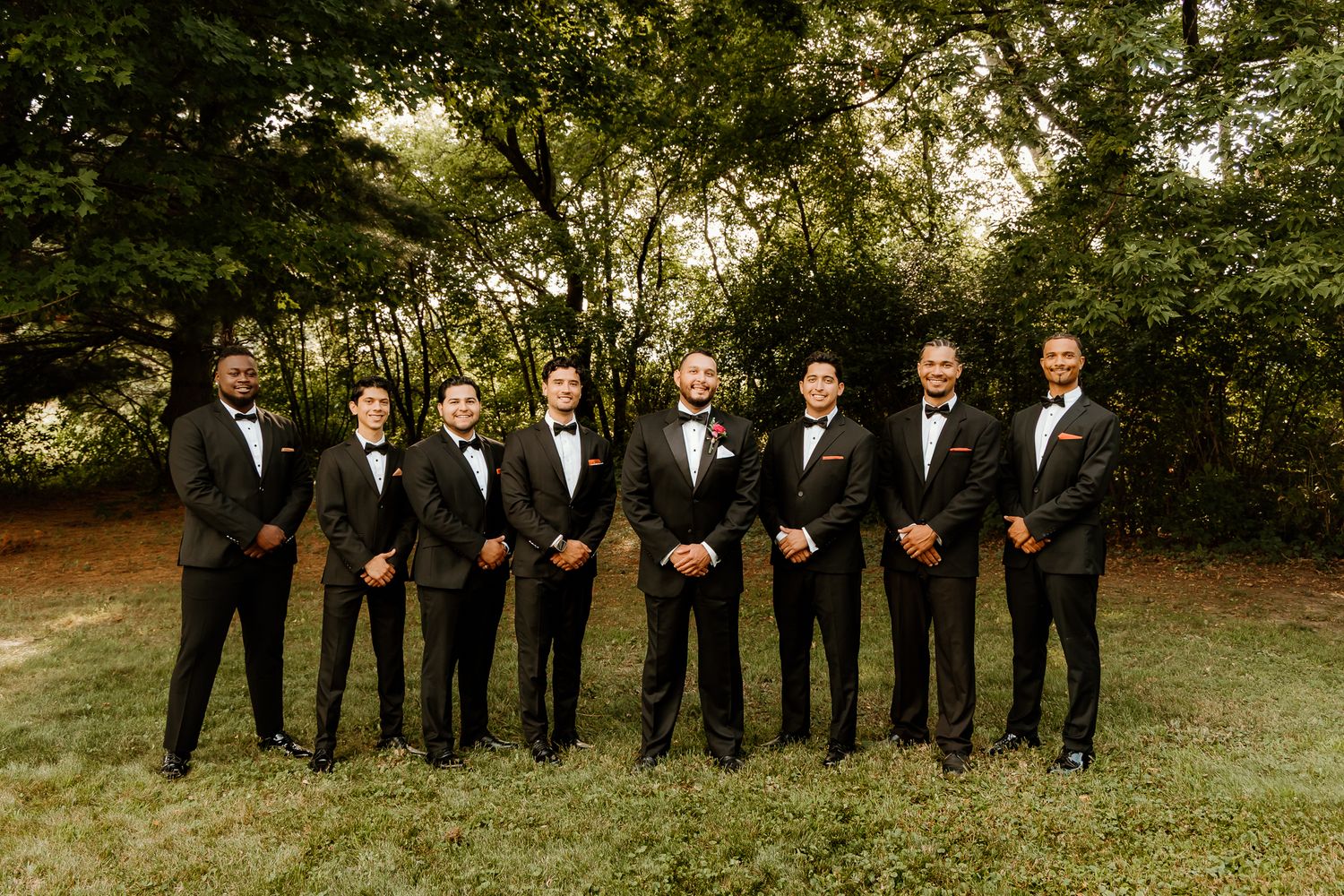 Groom and groomsmen in matching black tuxedos with bow ties posing outdoors on green grass with trees behind them.