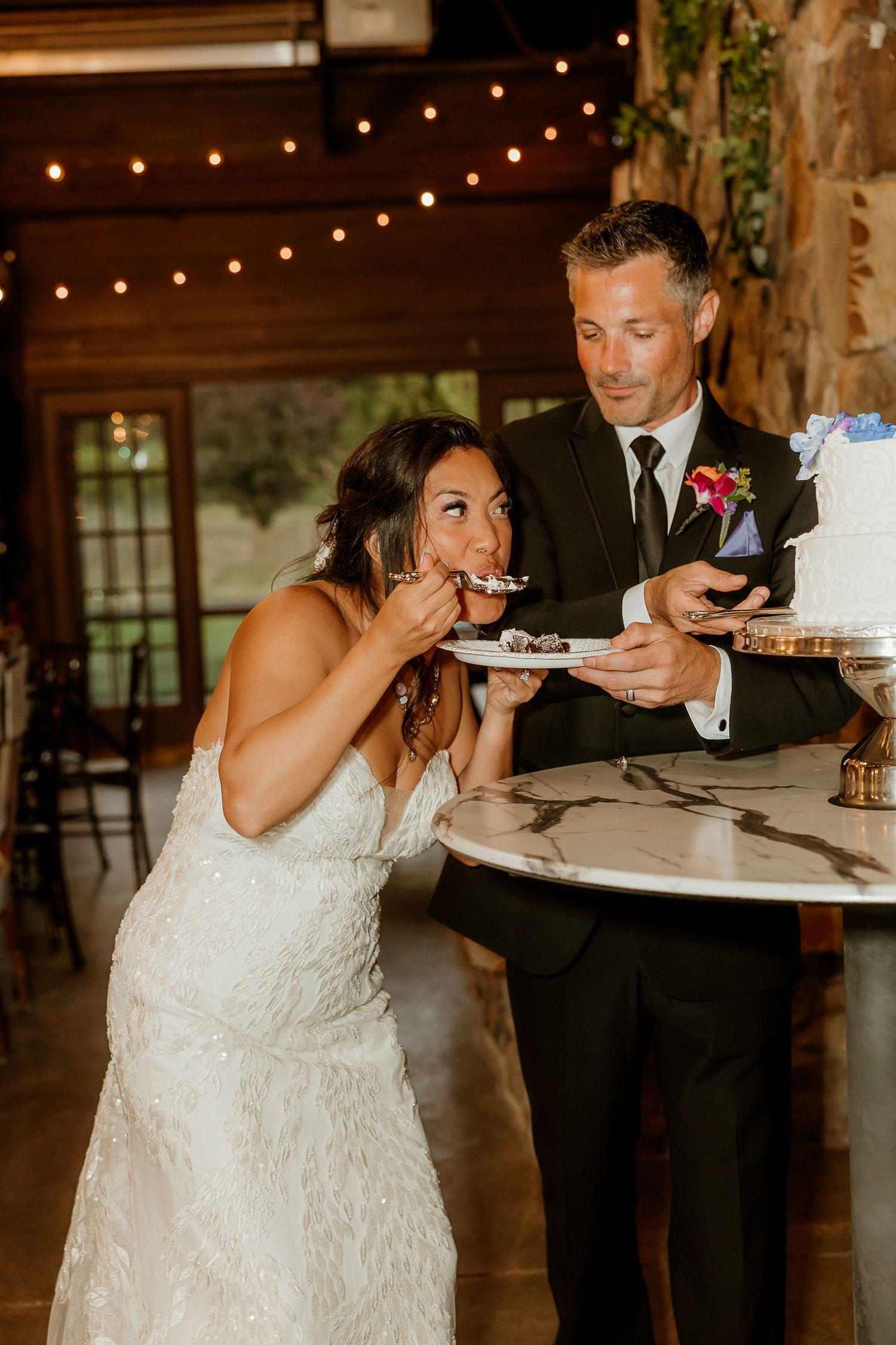 Bride in white gown eating wedding cake beside groom in black suit at rustic venue with string lights.