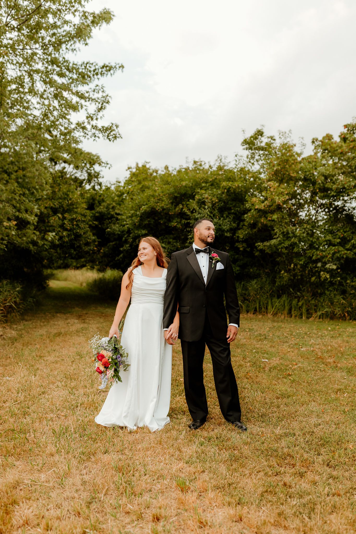A bride and groom hold hands in a green field, she in a white gown with bouquet, he in a black tuxedo.