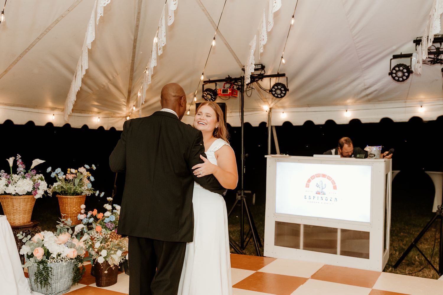 Couple sharing first dance on checkered floor under white tent at elegant wedding reception with DJ booth.