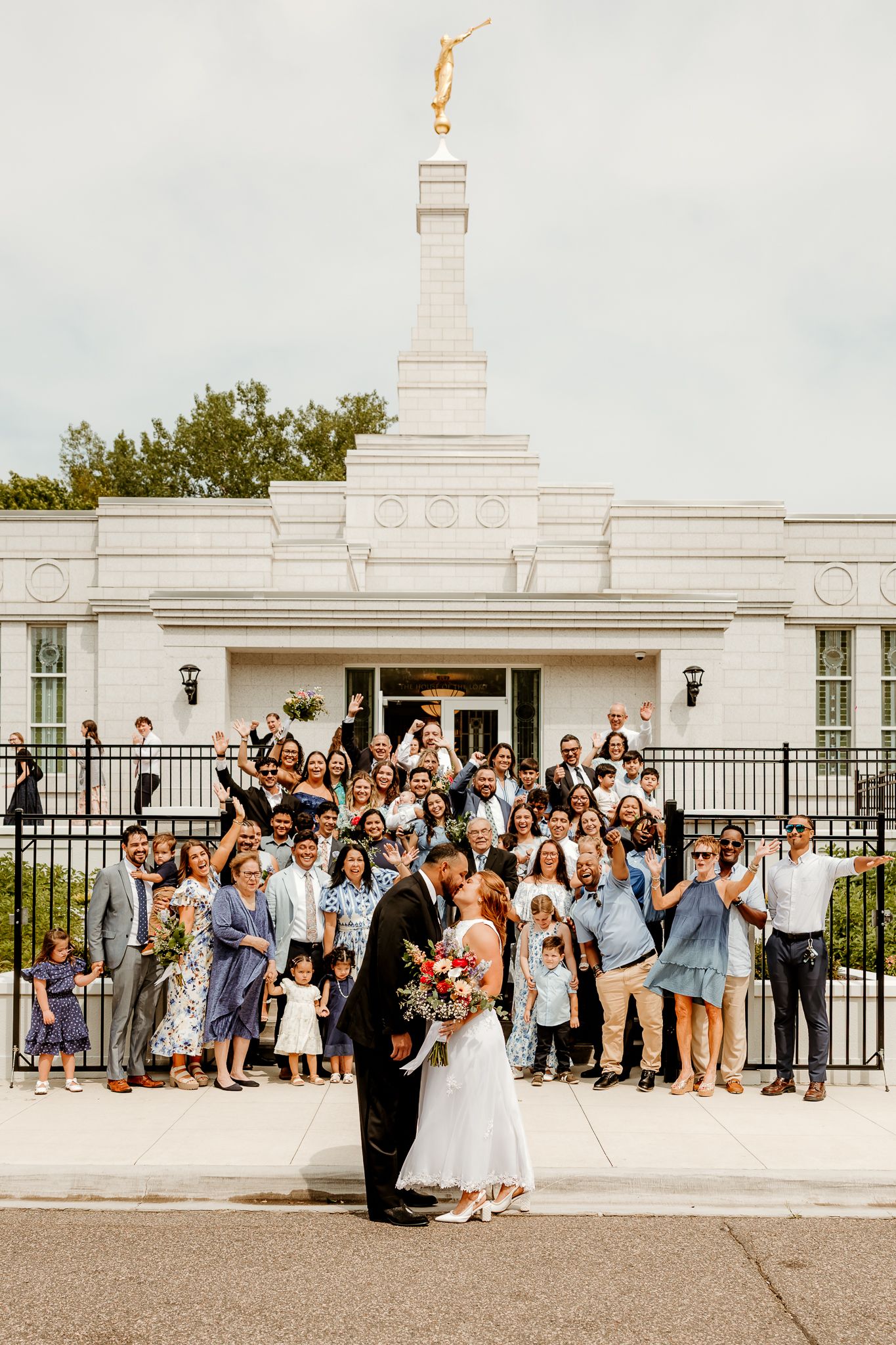 Newlywed couple kissing outside an LDS temple with cheering wedding guests gathered on the steps behind them.
