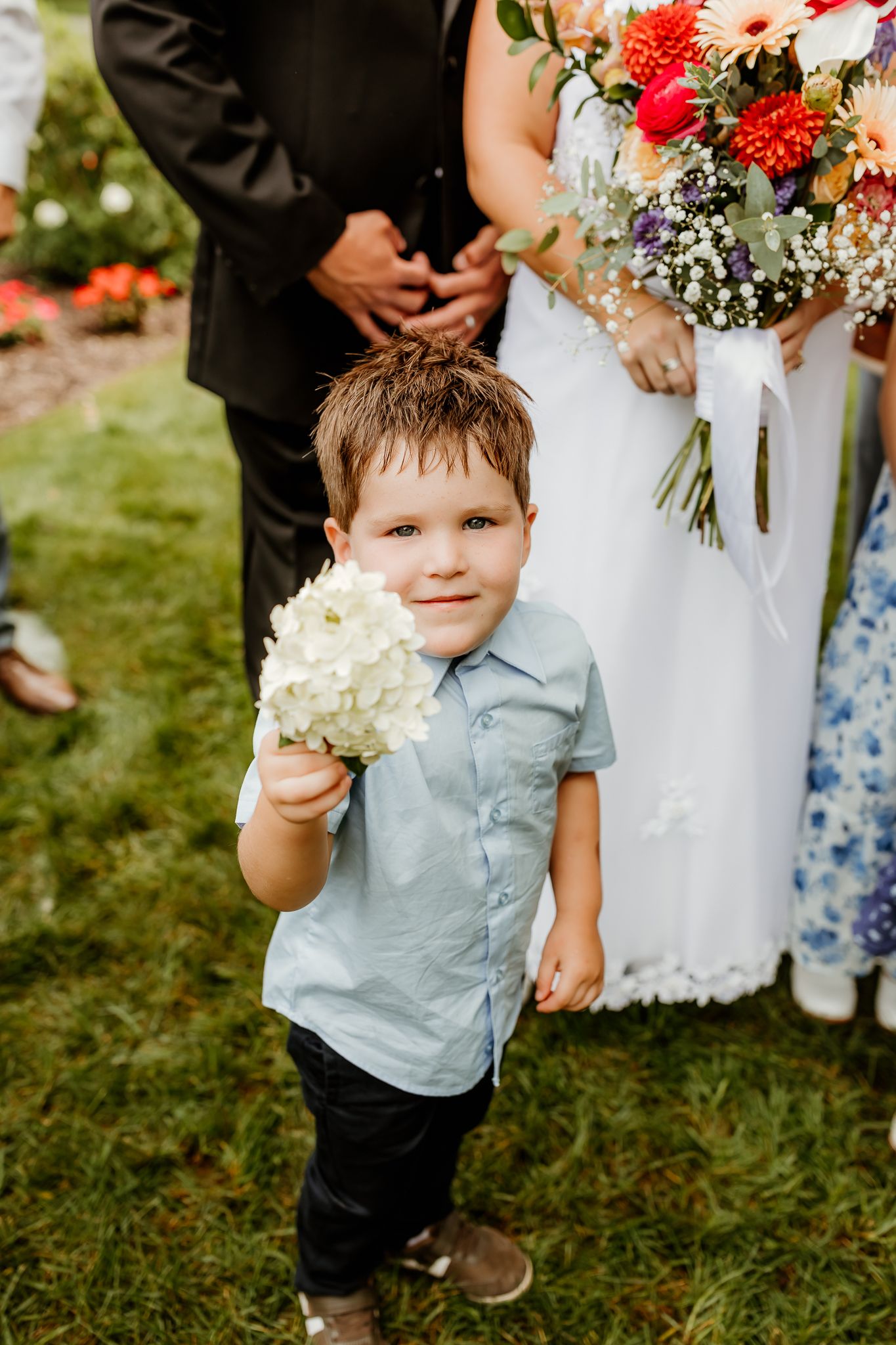 Young boy in blue shirt holds white hydrangea flowers at a wedding ceremony outdoors on green grass.